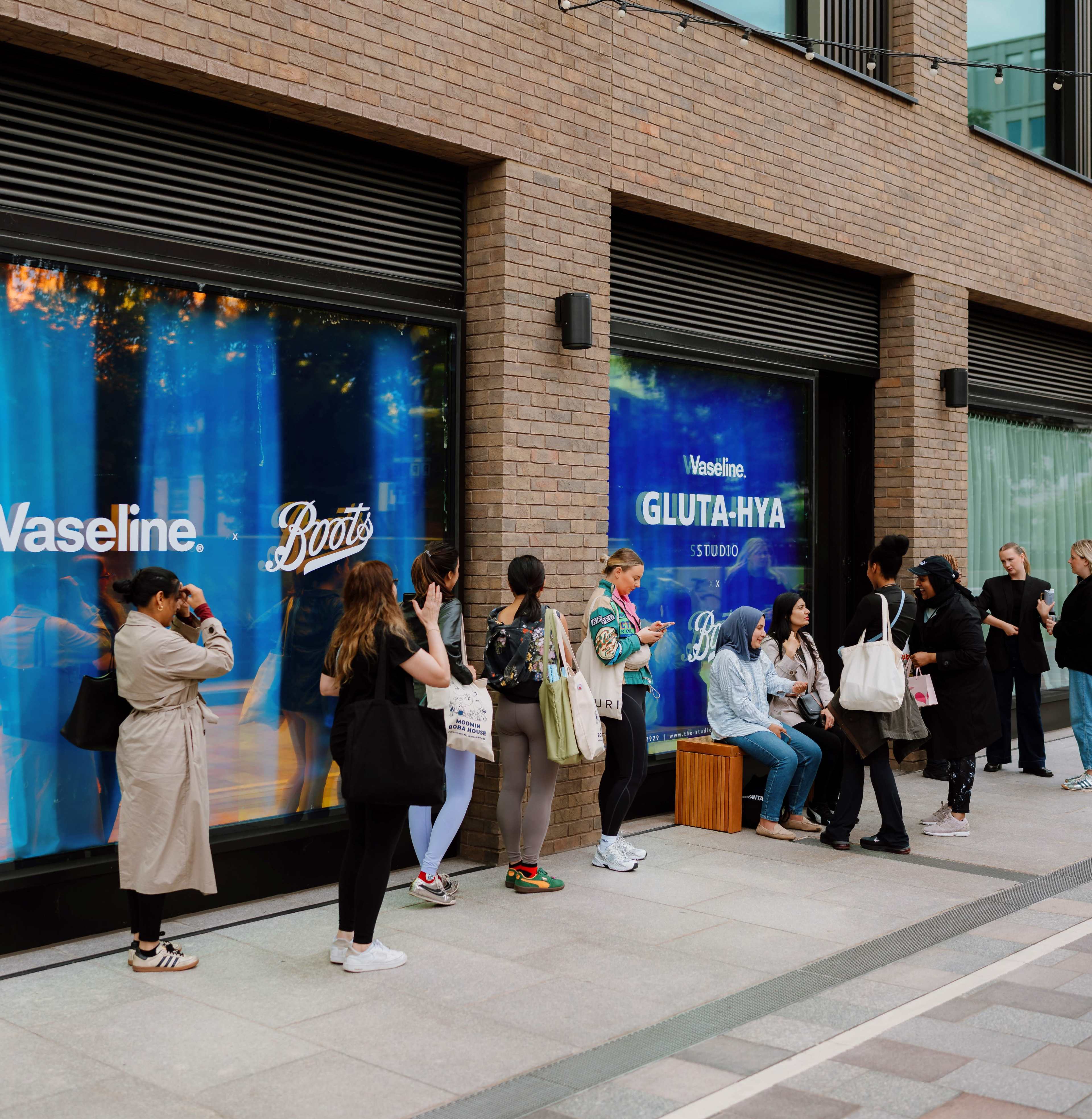 A group of people is lined up outside a storefront featuring advertisements for Vaseline and Boots.
