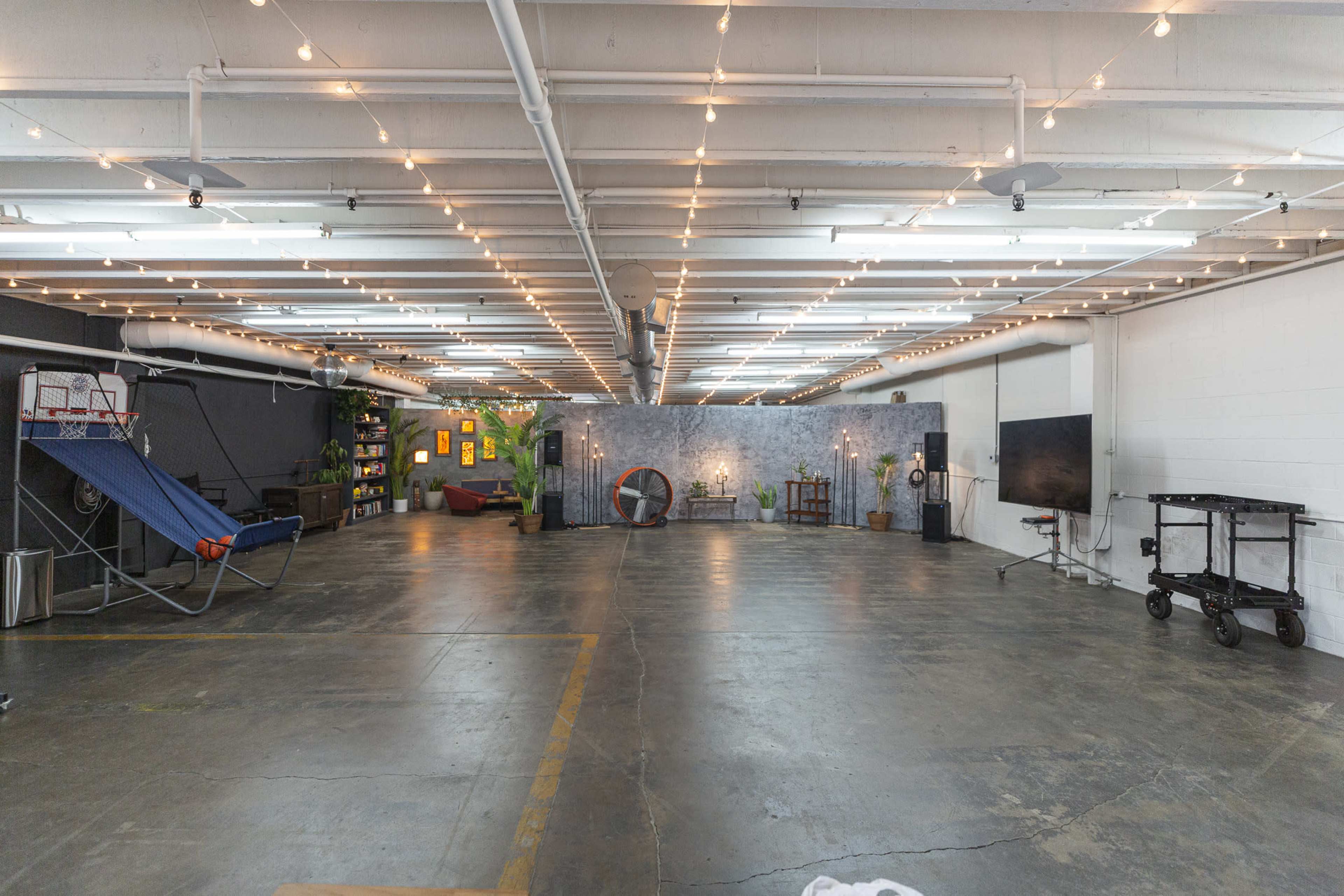 A spacious, industrial-style room with overhead lights, an open floor area, and various furnishings including a hammock, a television, and decorative plants.