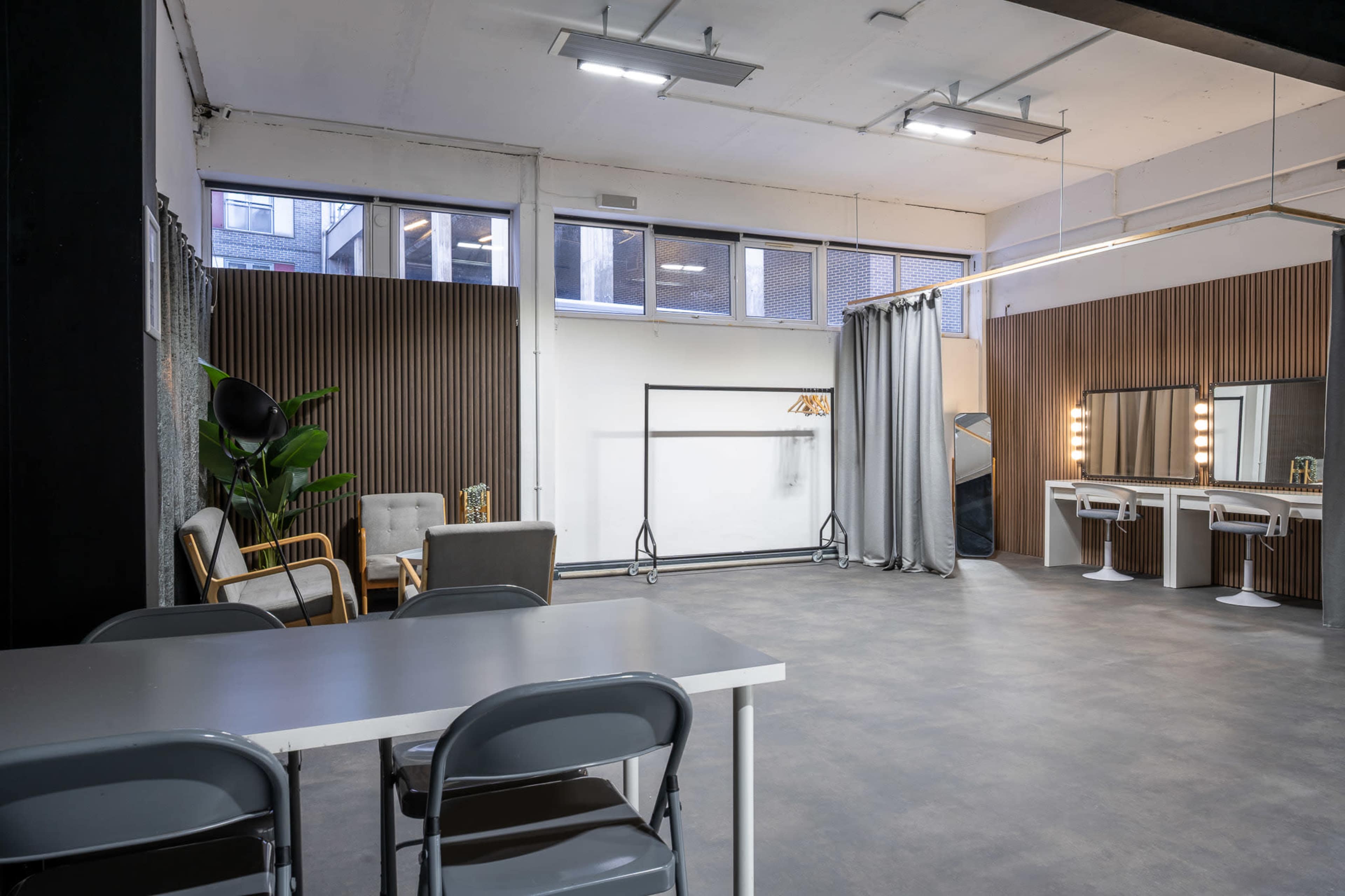 The image shows a spacious studio with a seating area, mirrors, and a clothing rack, all set against a backdrop of large windows and wooden paneling.