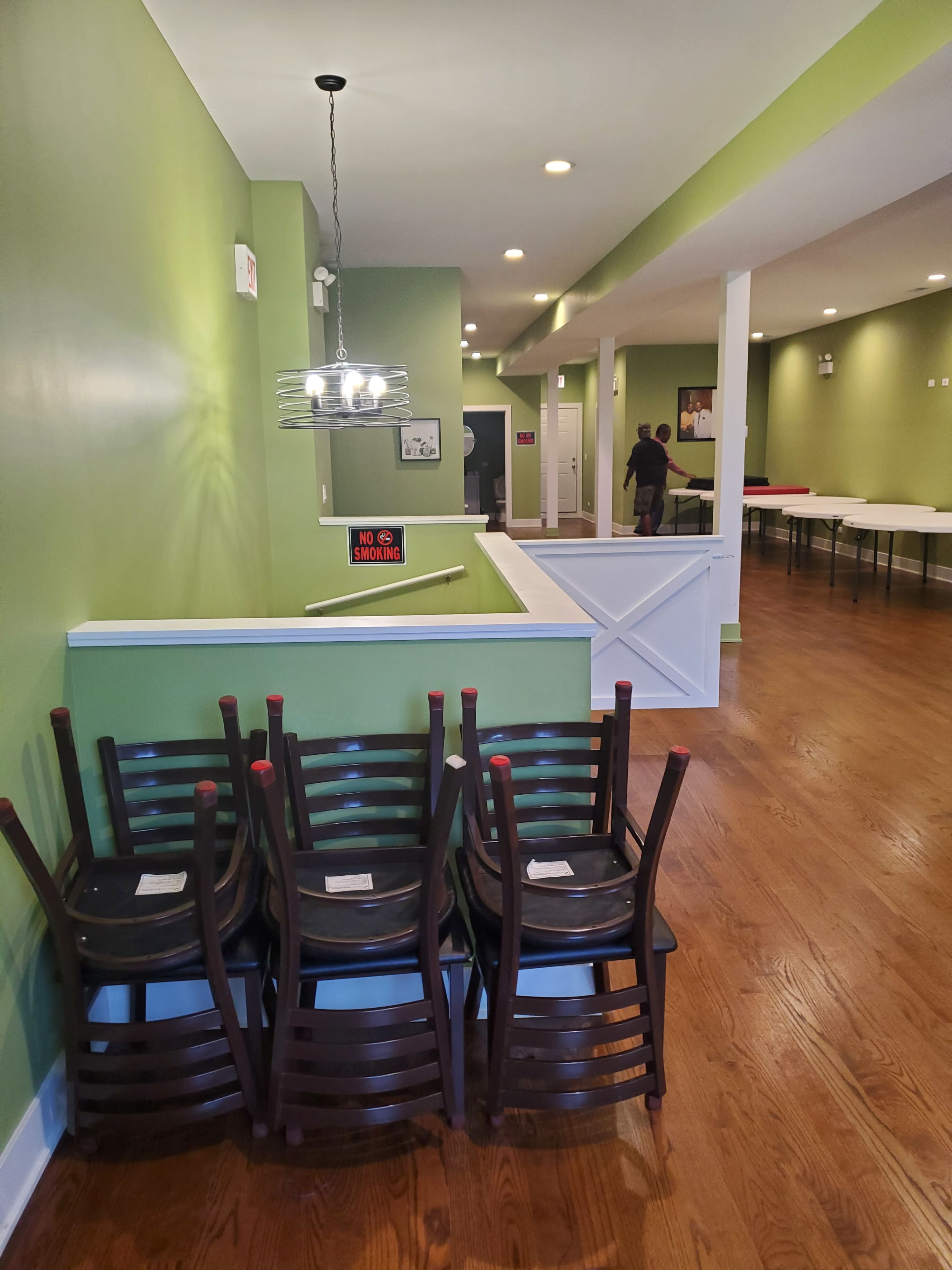 The interior of a restaurant features green walls, wooden flooring, and a stack of chairs against the wall, with tables set up in the background.