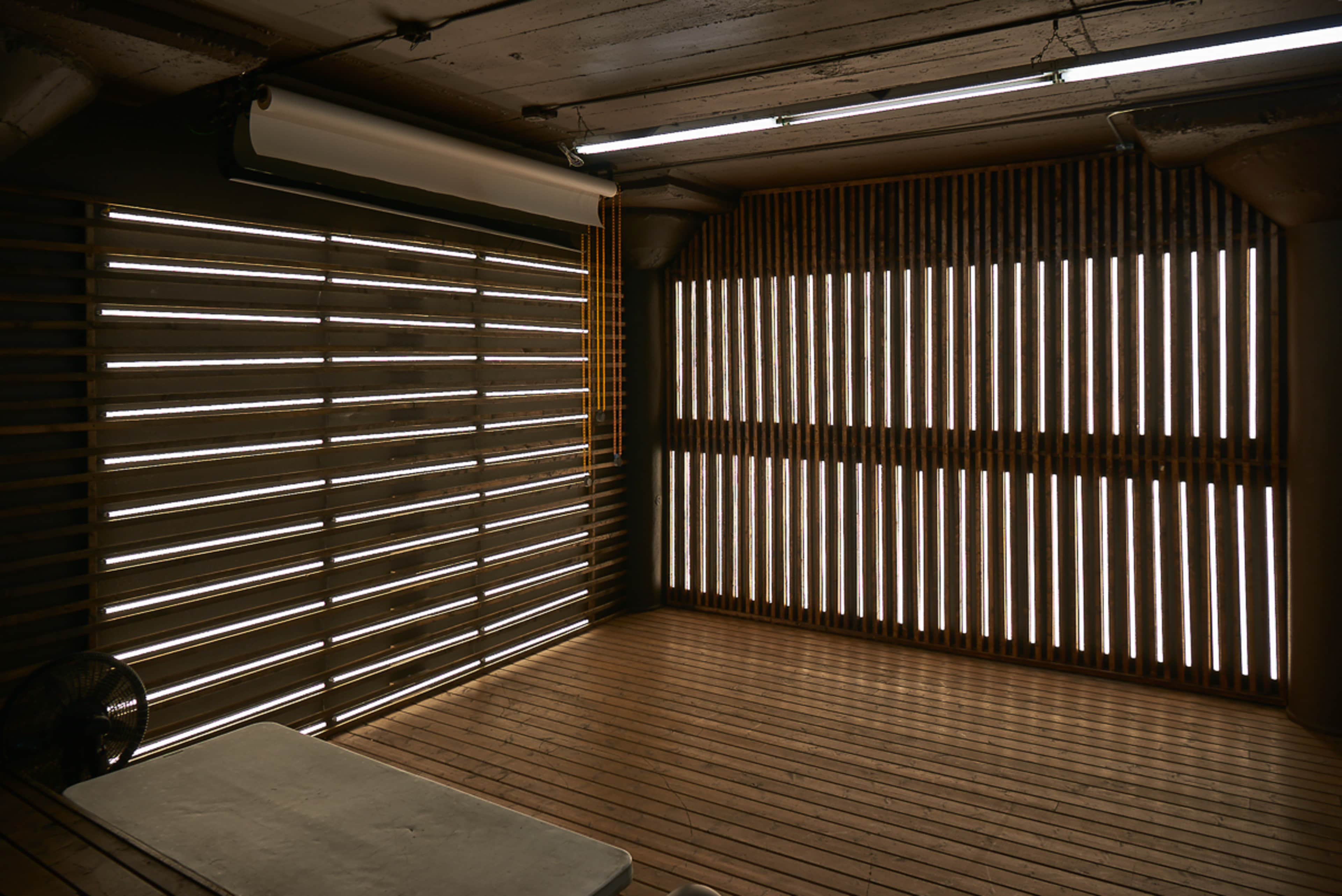 A minimalist interior space with wooden slatted walls and flooring, illuminated by natural light filtering through the slats.