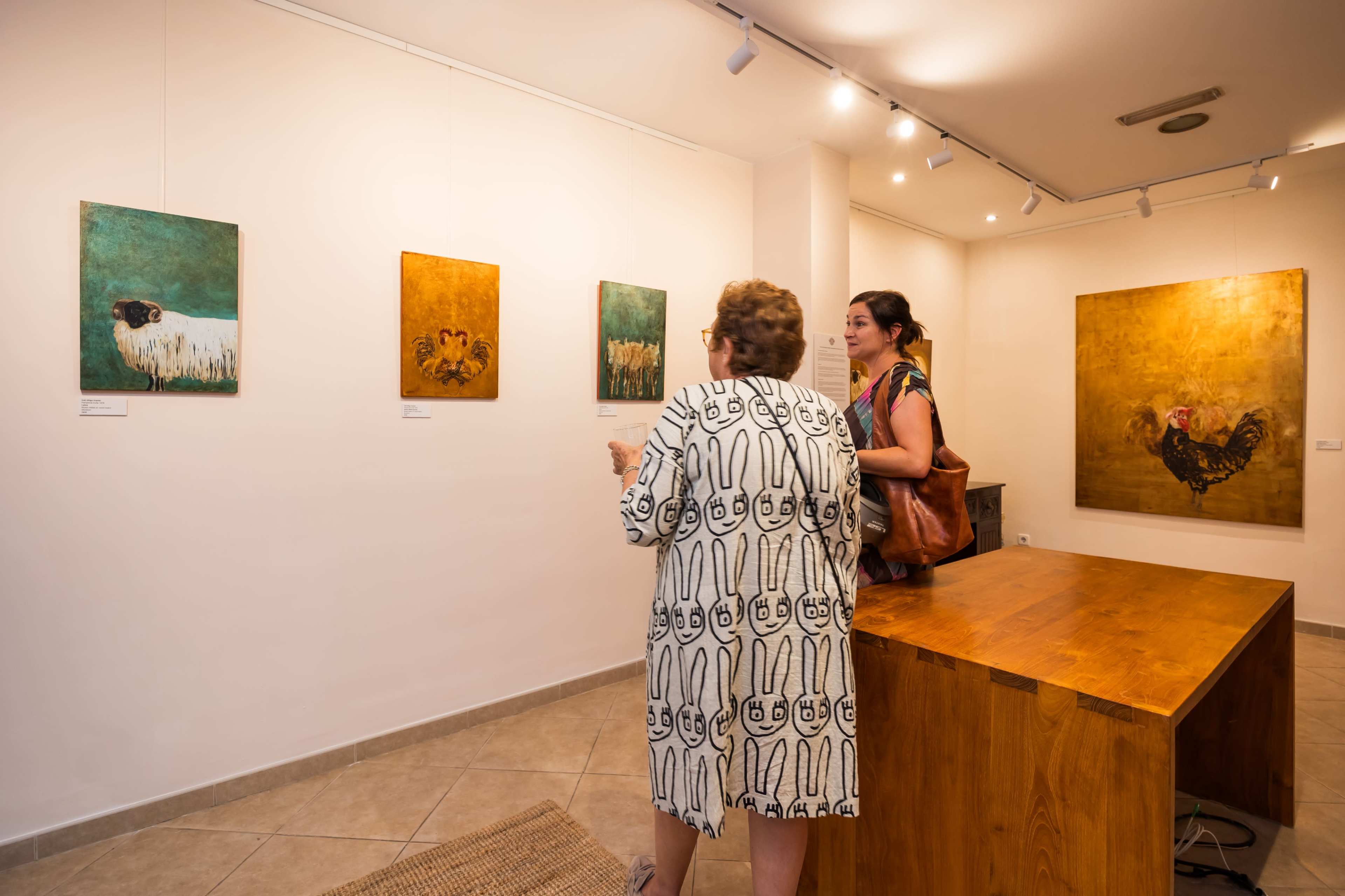 Two women are viewing art pieces in an exhibition space, with paintings displayed on white walls and a wooden table in the foreground.