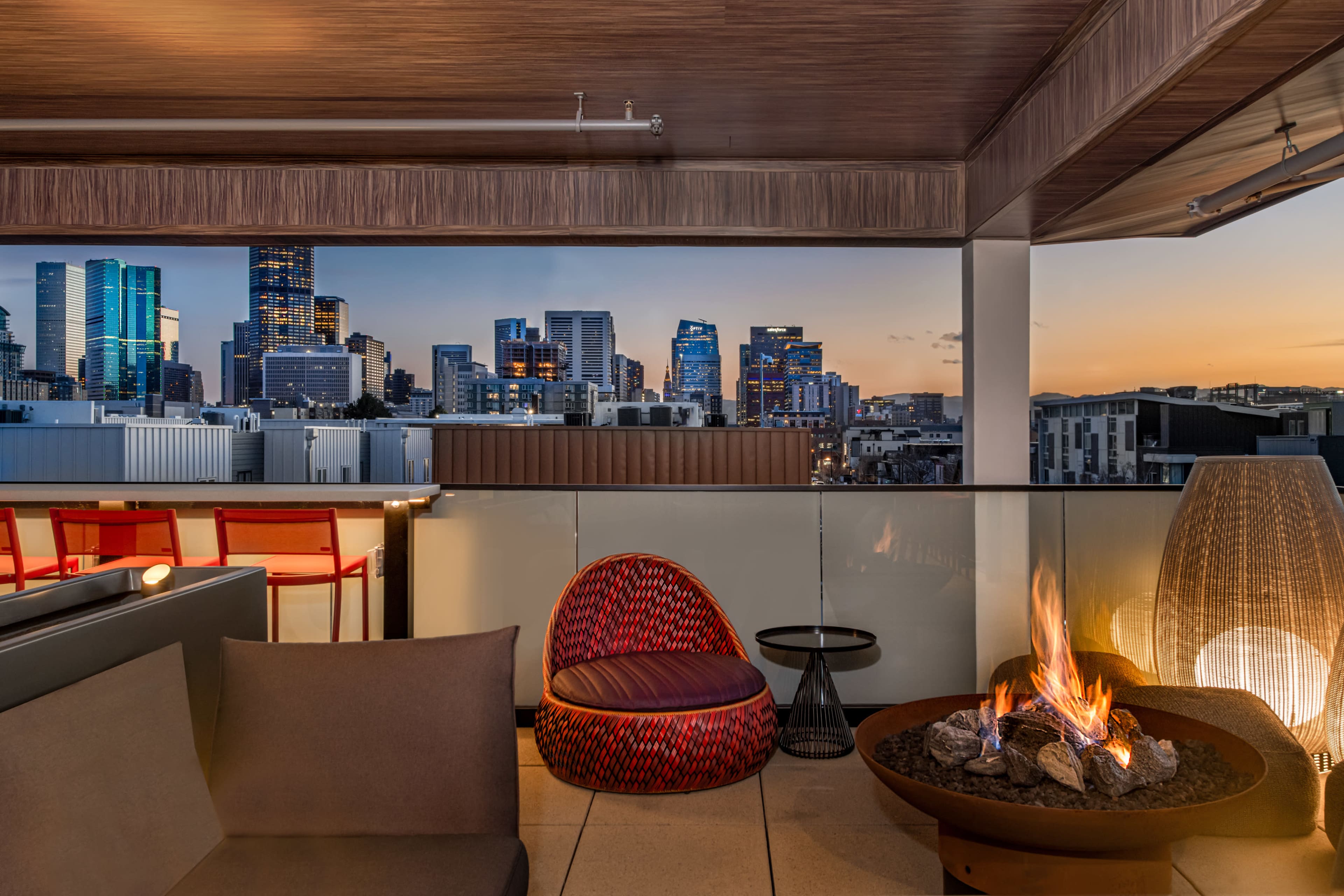 A rooftop lounge area with modern seating and a fire pit, overlooking a city skyline at sunset.