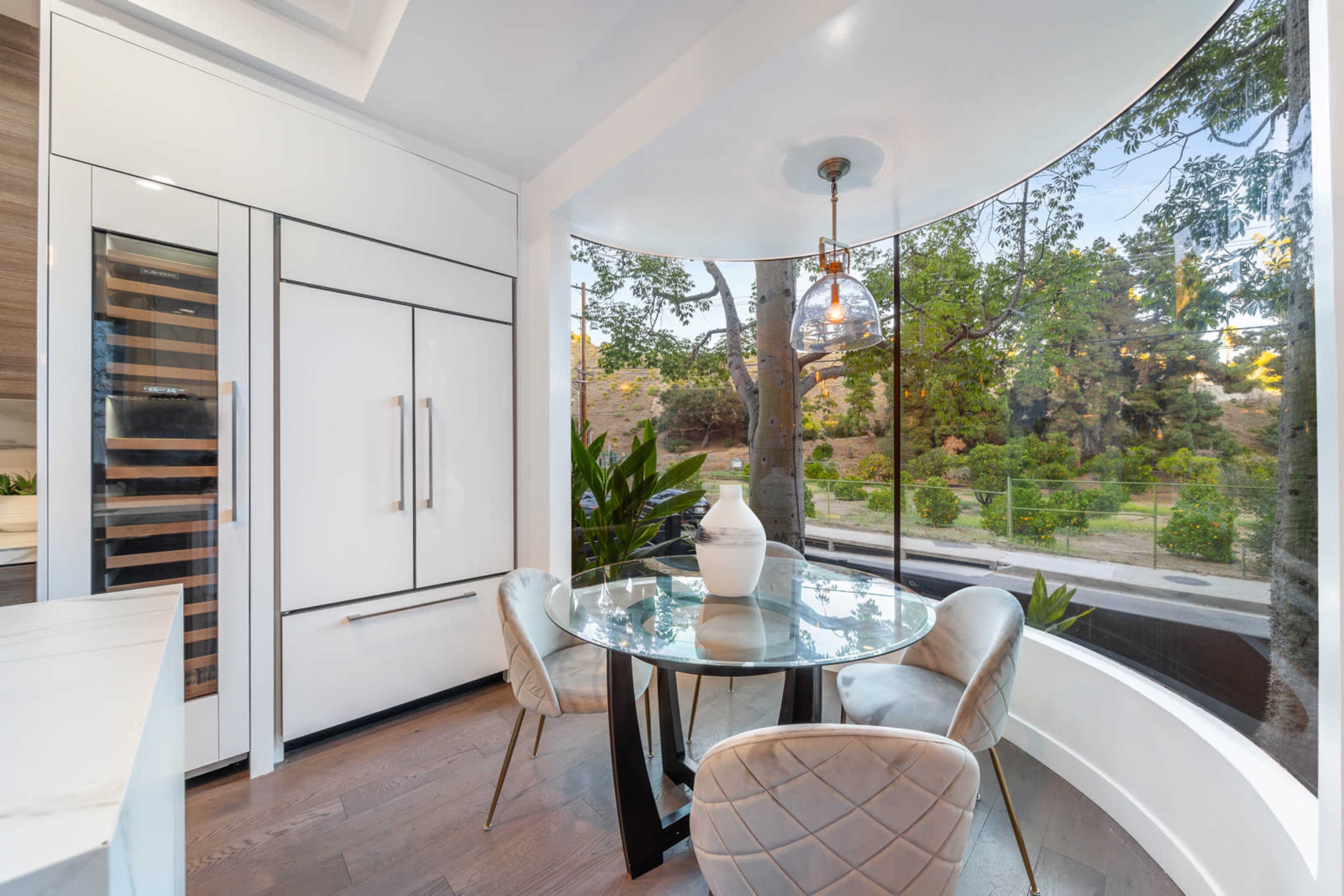 A round glass table and four chairs are set in a bright corner dining area with large windows overlooking a garden.