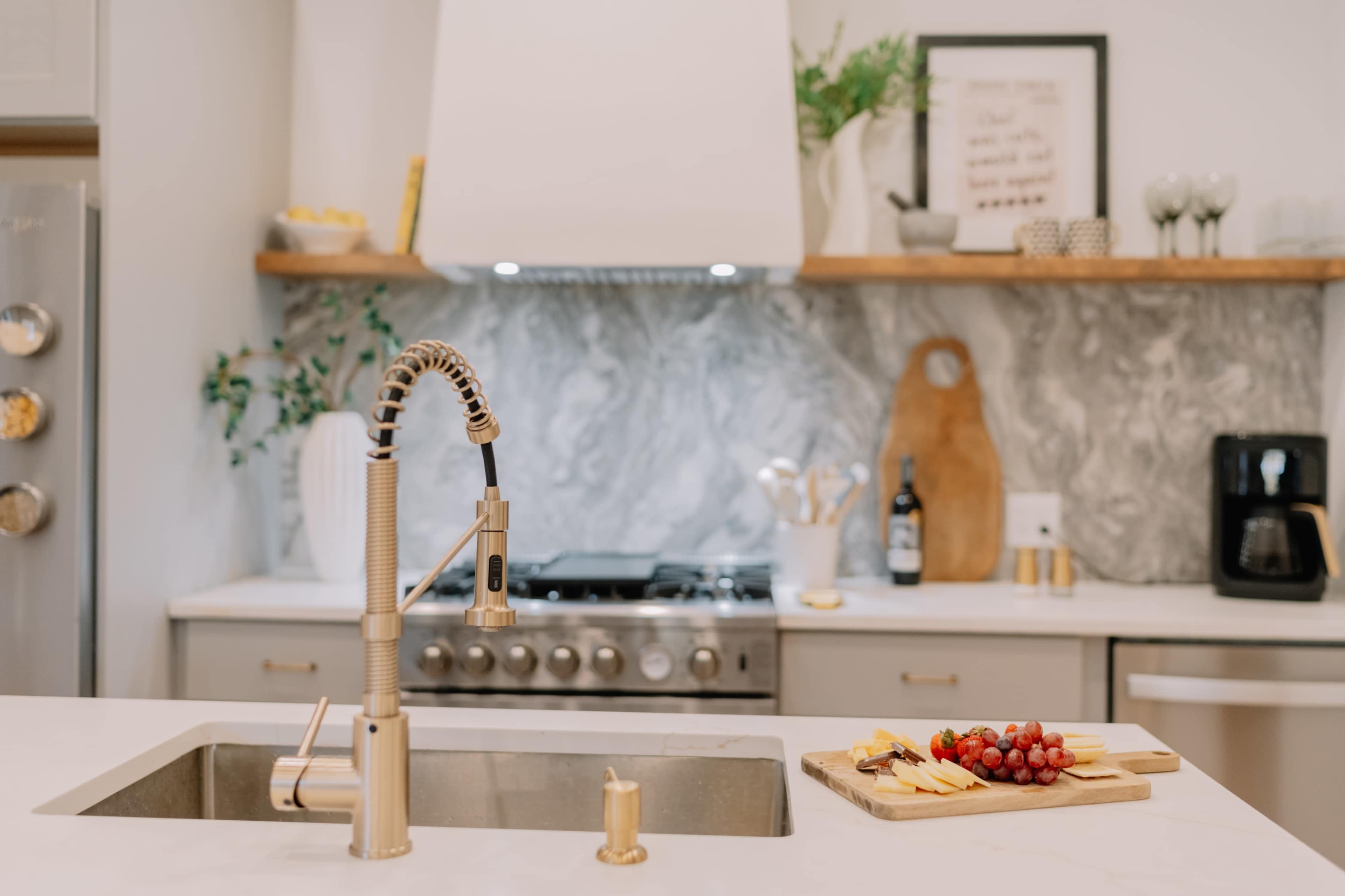 A modern kitchen features a stainless steel sink with a gold faucet, adjacent to a wooden cutting board with cheese and grapes.