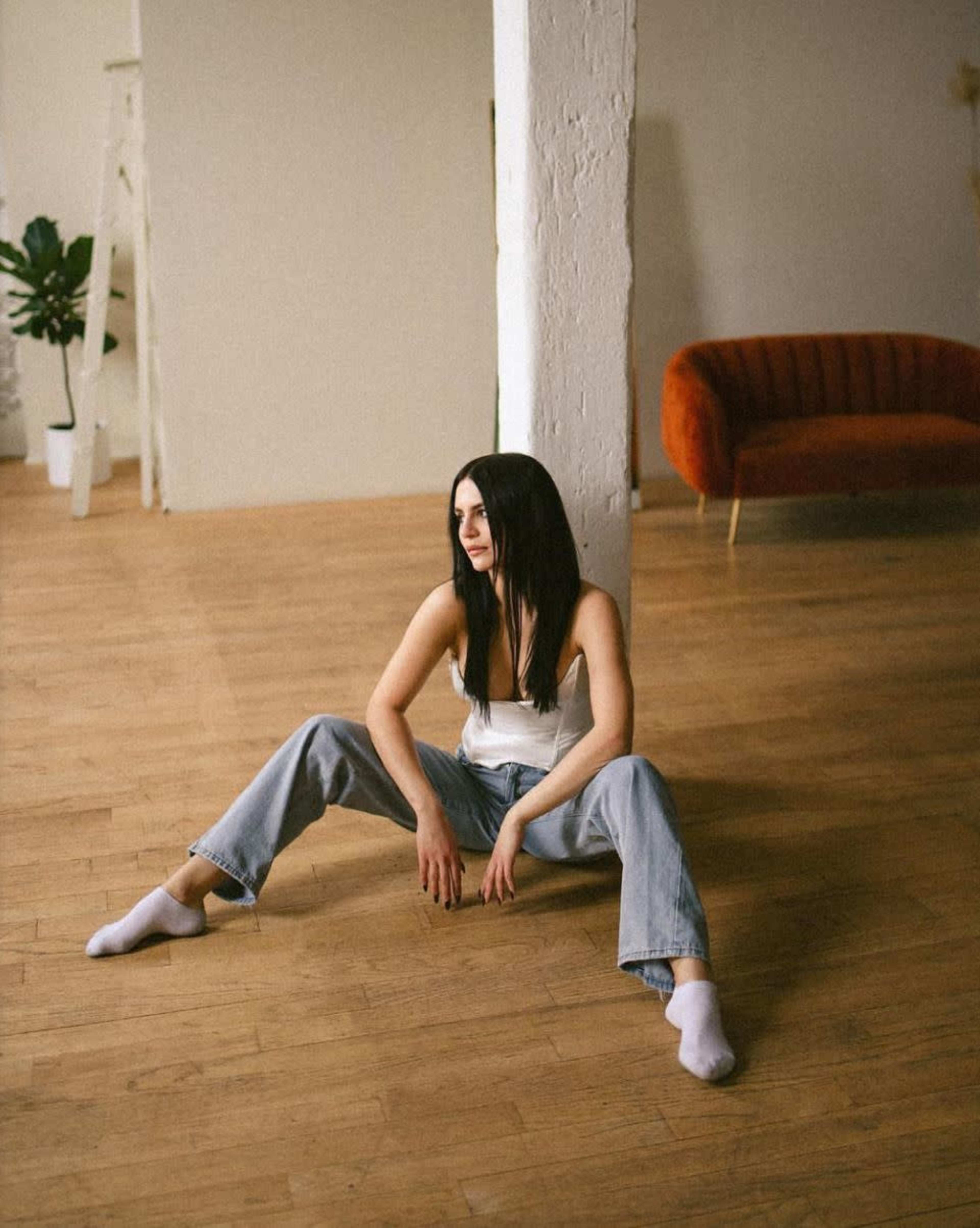 A woman sits on a wooden floor with her legs spread apart, leaning against a white column in a spacious room featuring a cozy sofa and a plant.