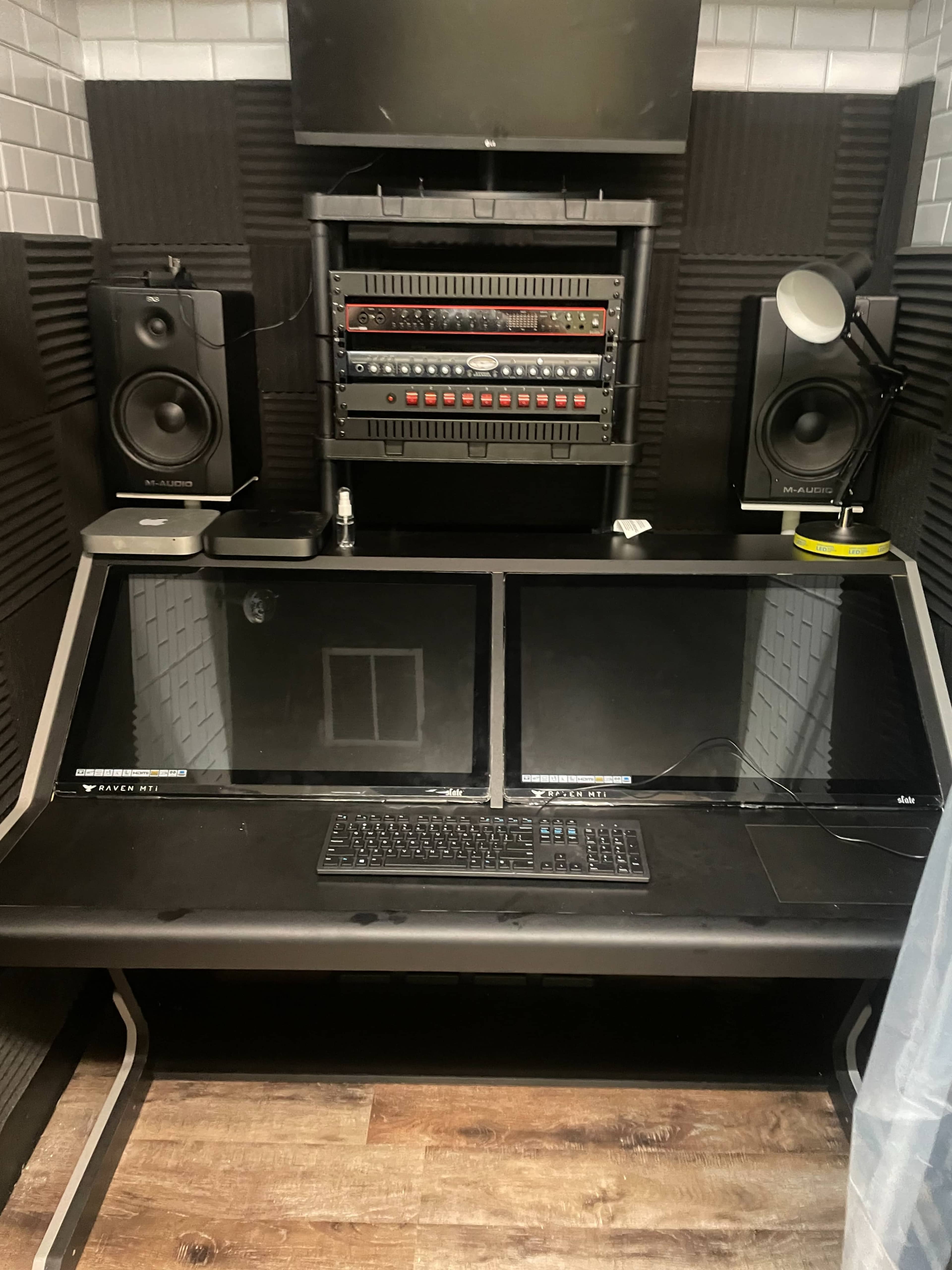 A professional recording studio setup with a double-monitor desk, speakers on either side, and audio equipment mounted above.