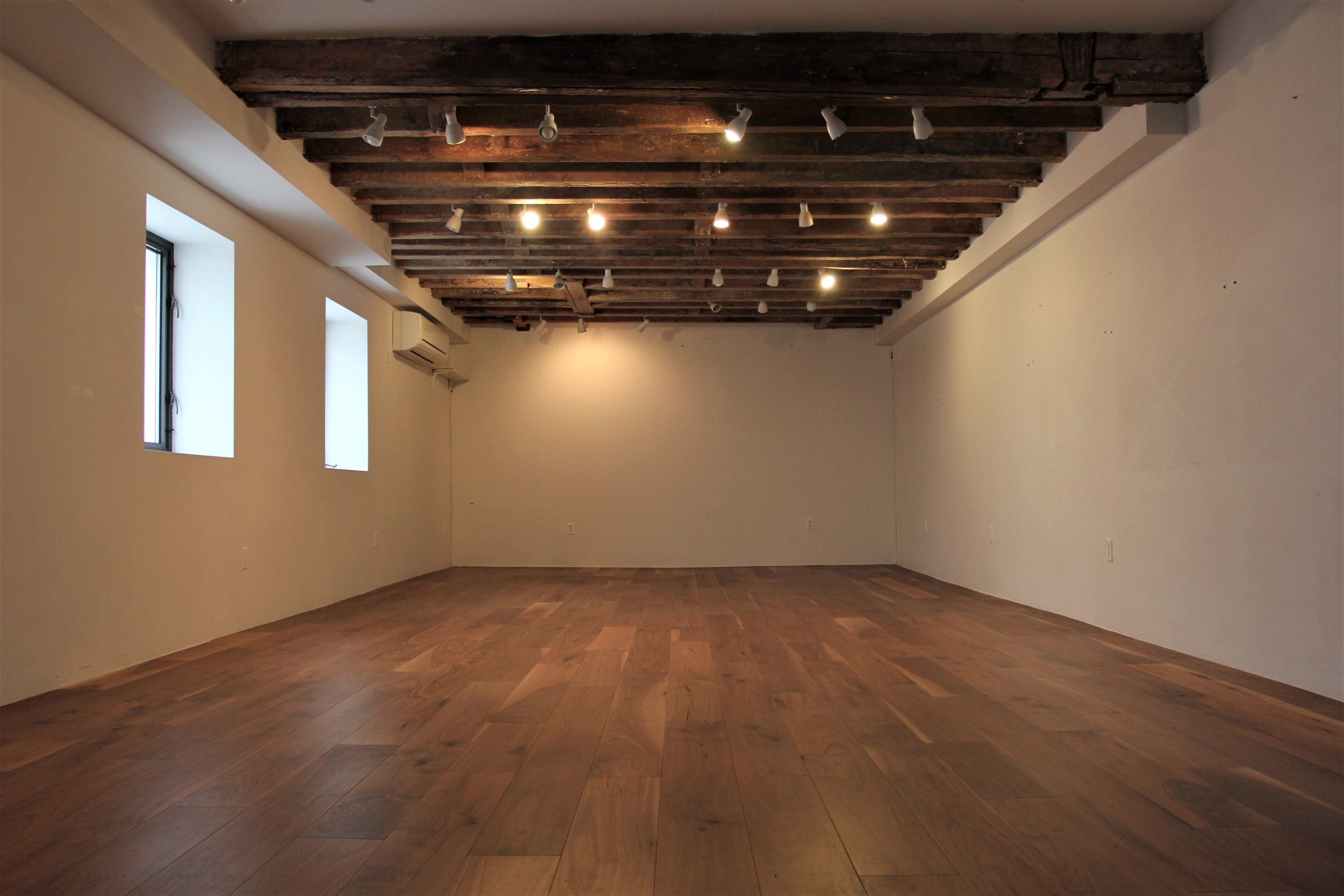 An empty room with wooden floors, exposed beams on the ceiling, and two windows letting in natural light.