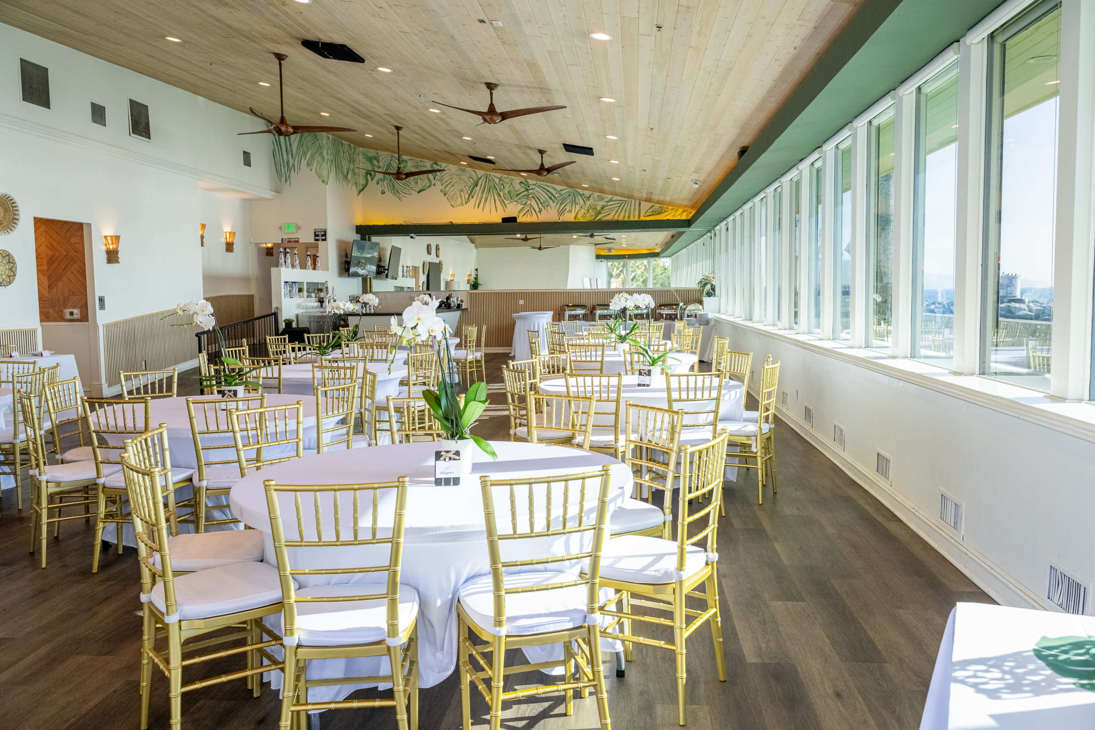 The interior features a spacious event venue with rows of round tables set with white tablecloths and gold chairs, illuminated by natural light from large windows.