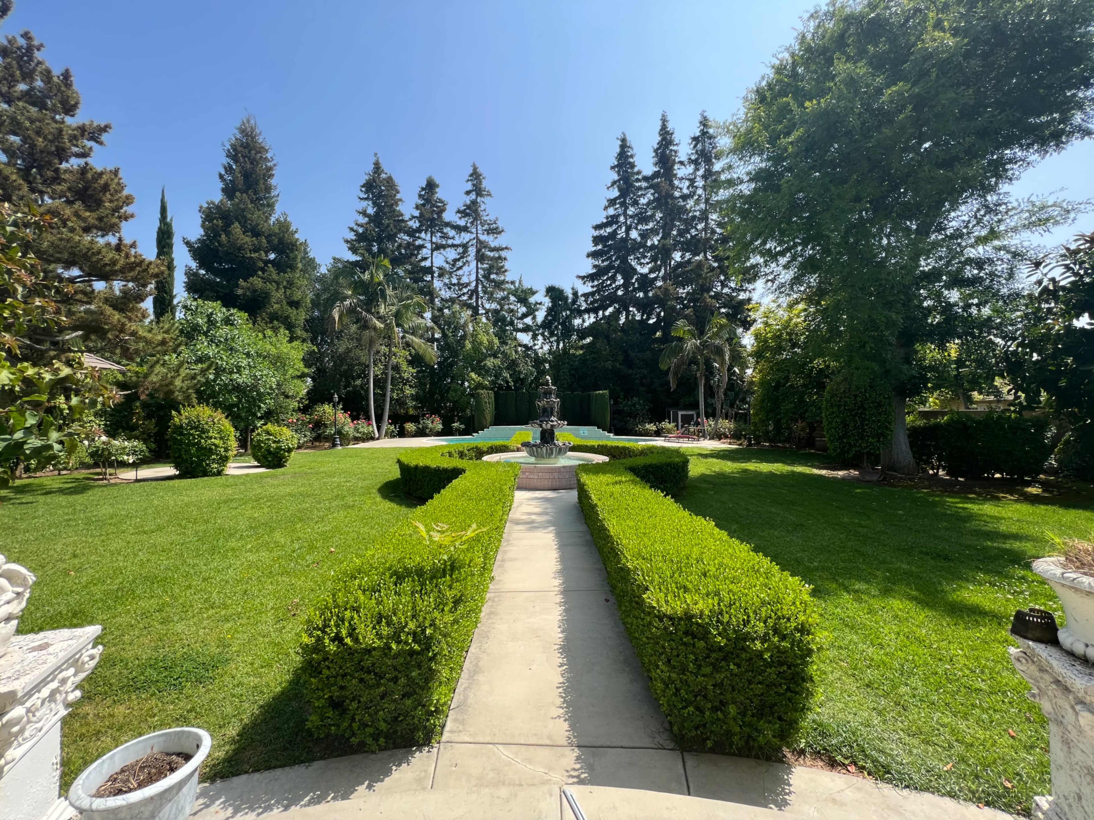 A well-manicured garden features neatly trimmed hedges, a stone pathway, and a fountain at the center, surrounded by tall trees and diverse greenery.