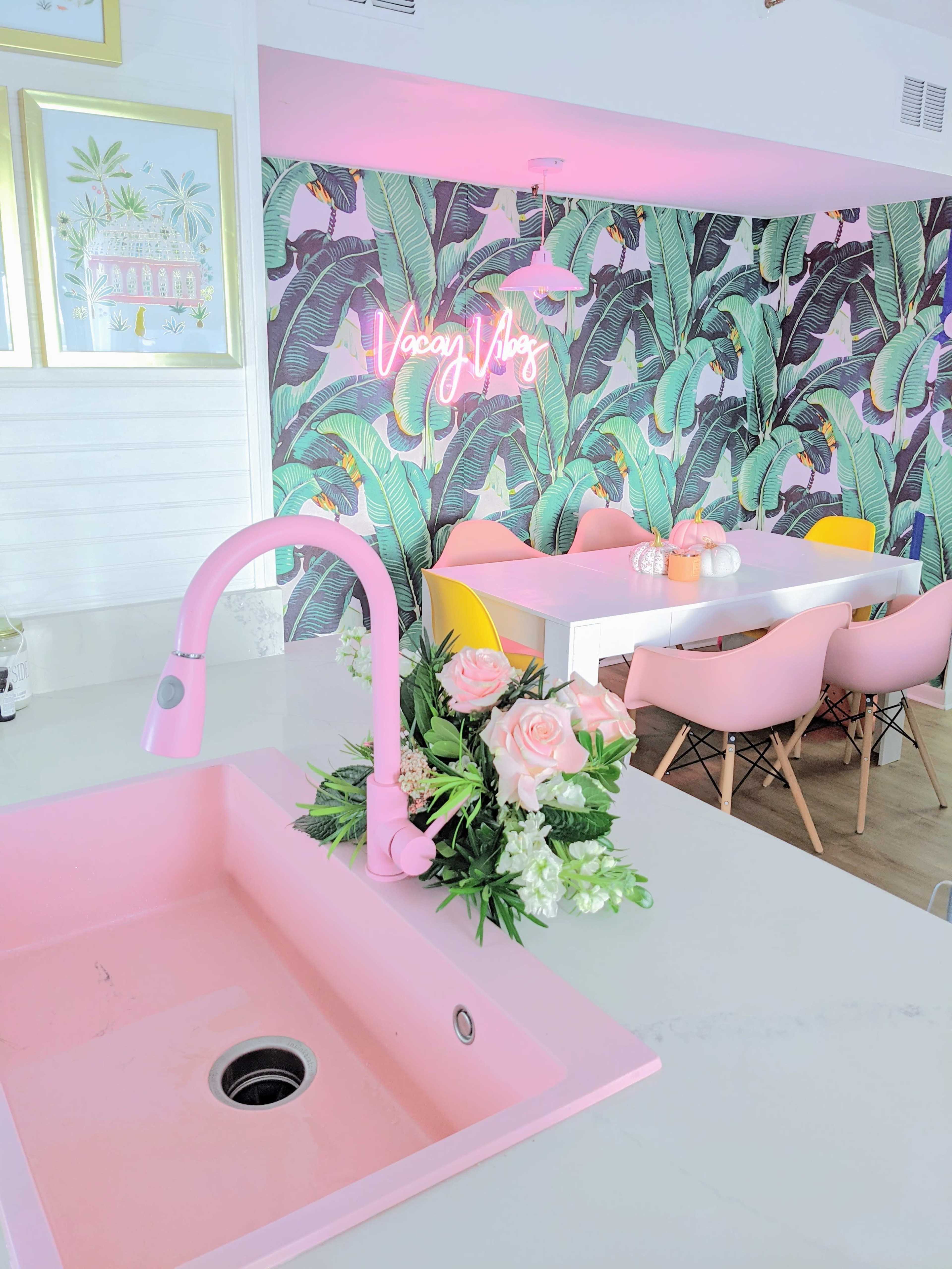 A modern kitchen features a pink sink and faucet, complemented by tropical wallpaper and a dining area with colorful chairs and a neon sign.