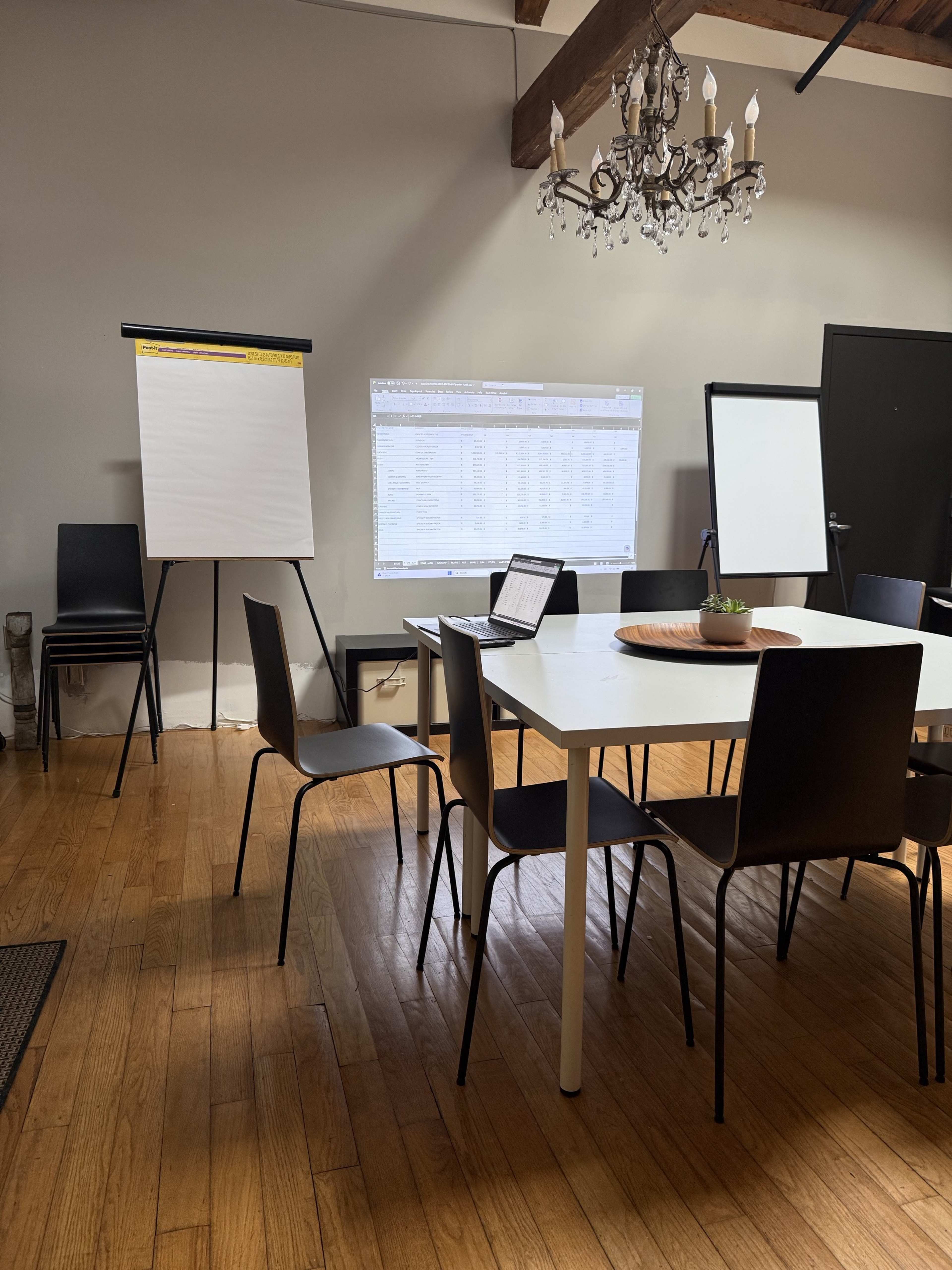 A modern meeting room features a round table surrounded by black chairs, with a laptop on the table and a presentation projected on the wall.