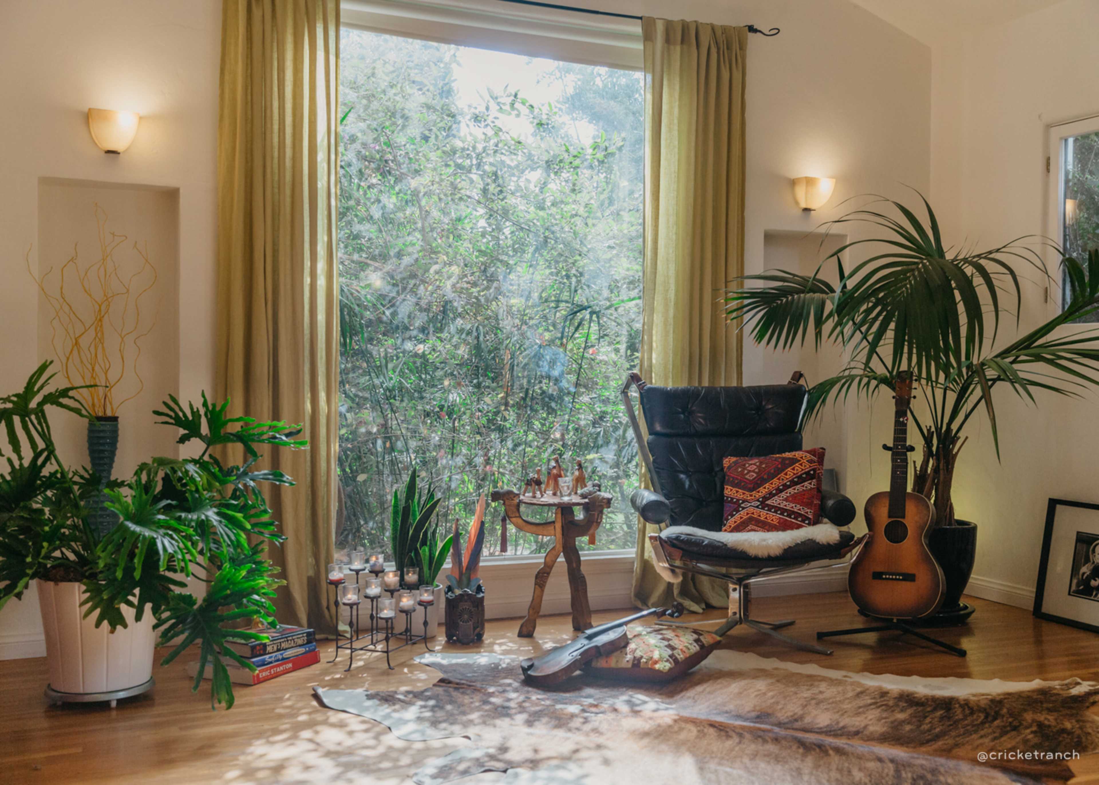 A cozy living room features a black armchair, a wooden table with decorative items, a guitar, and large potted plants beside a window draped with yellow curtains.