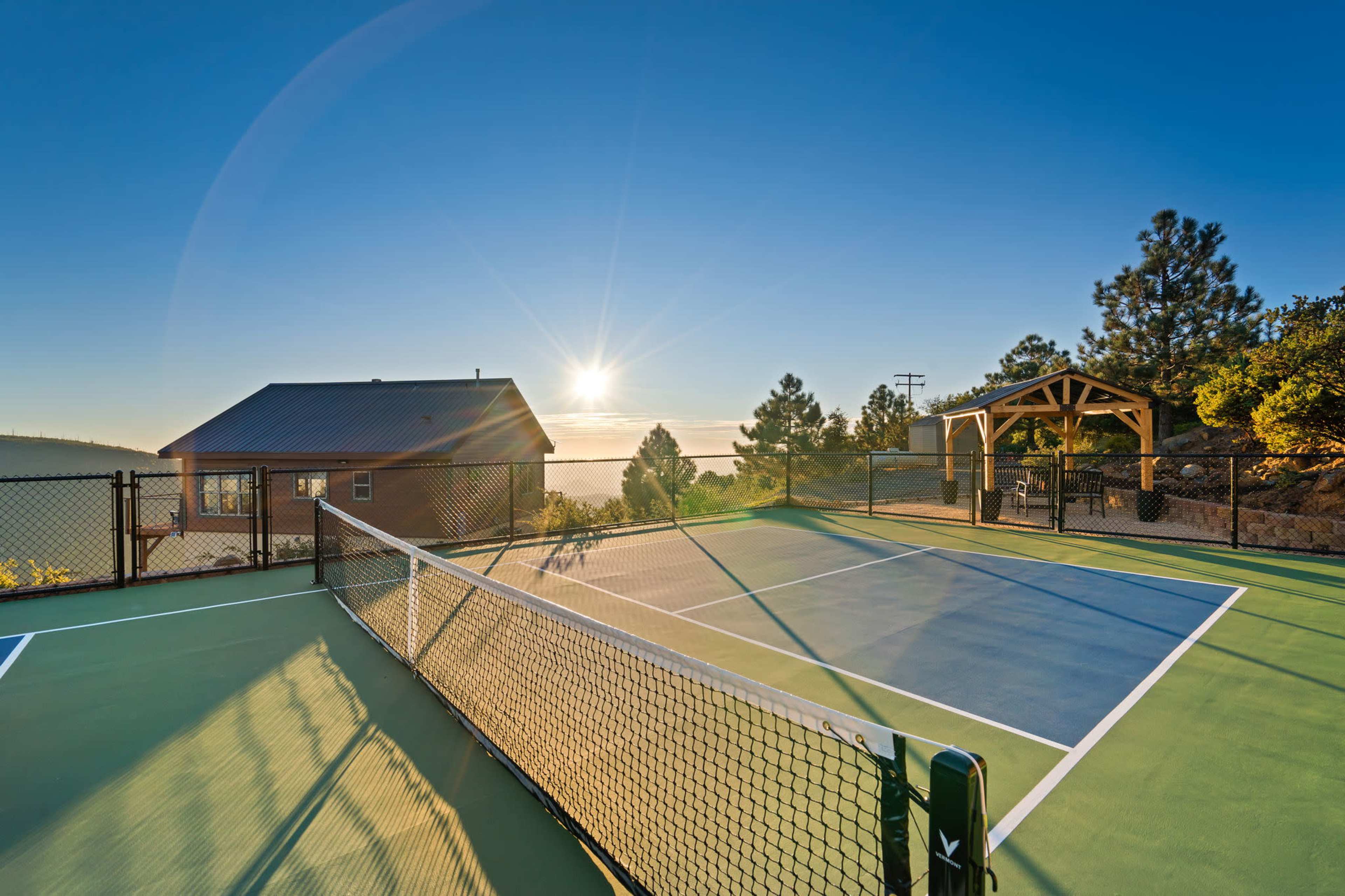 The image shows a tennis court adjacent to a wooden structure and a house, with a sunrise illuminating the scene.