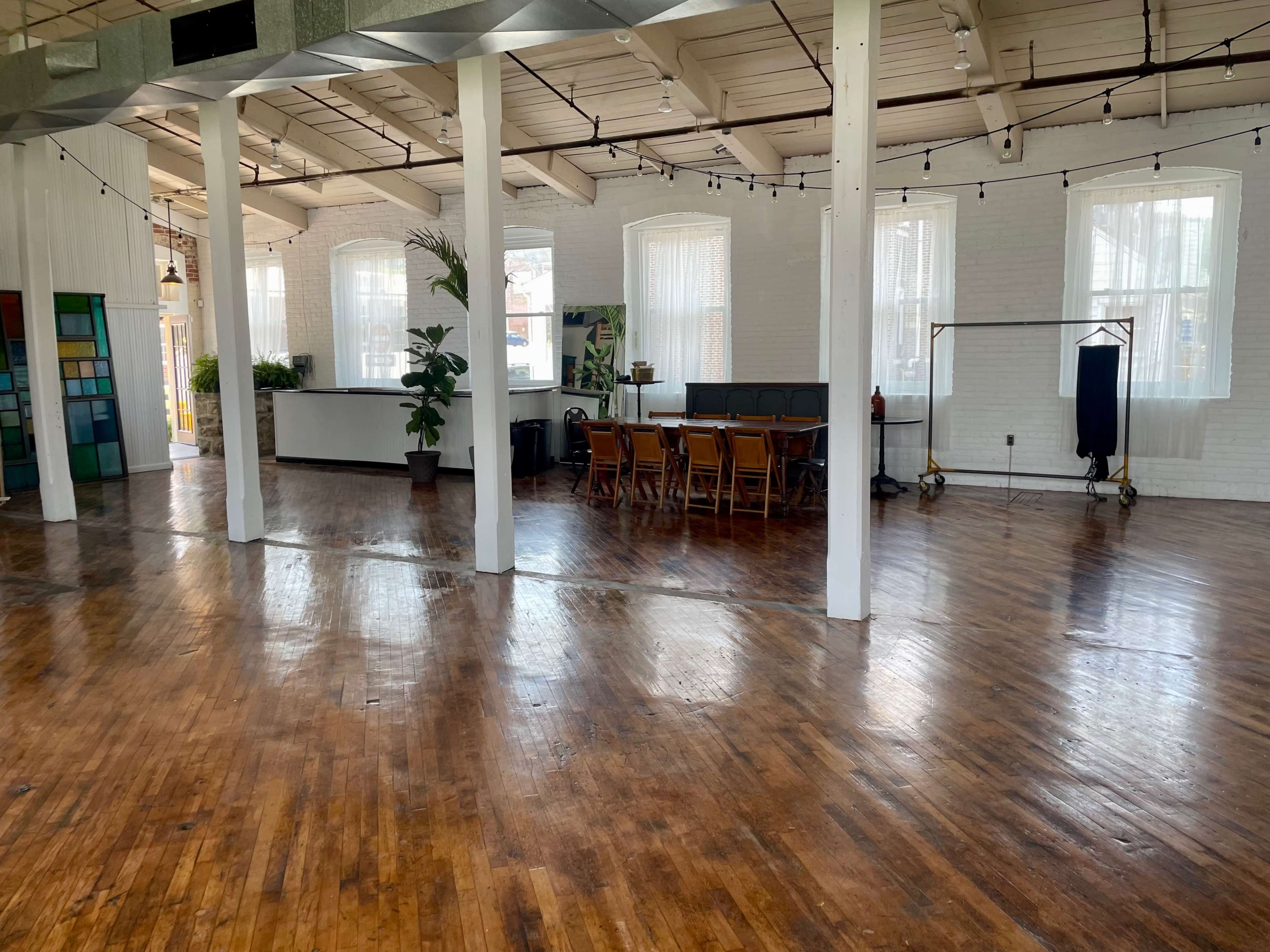 A spacious, bright room with wooden floors, exposed beams, large windows, a long dining table, and several potted plants.