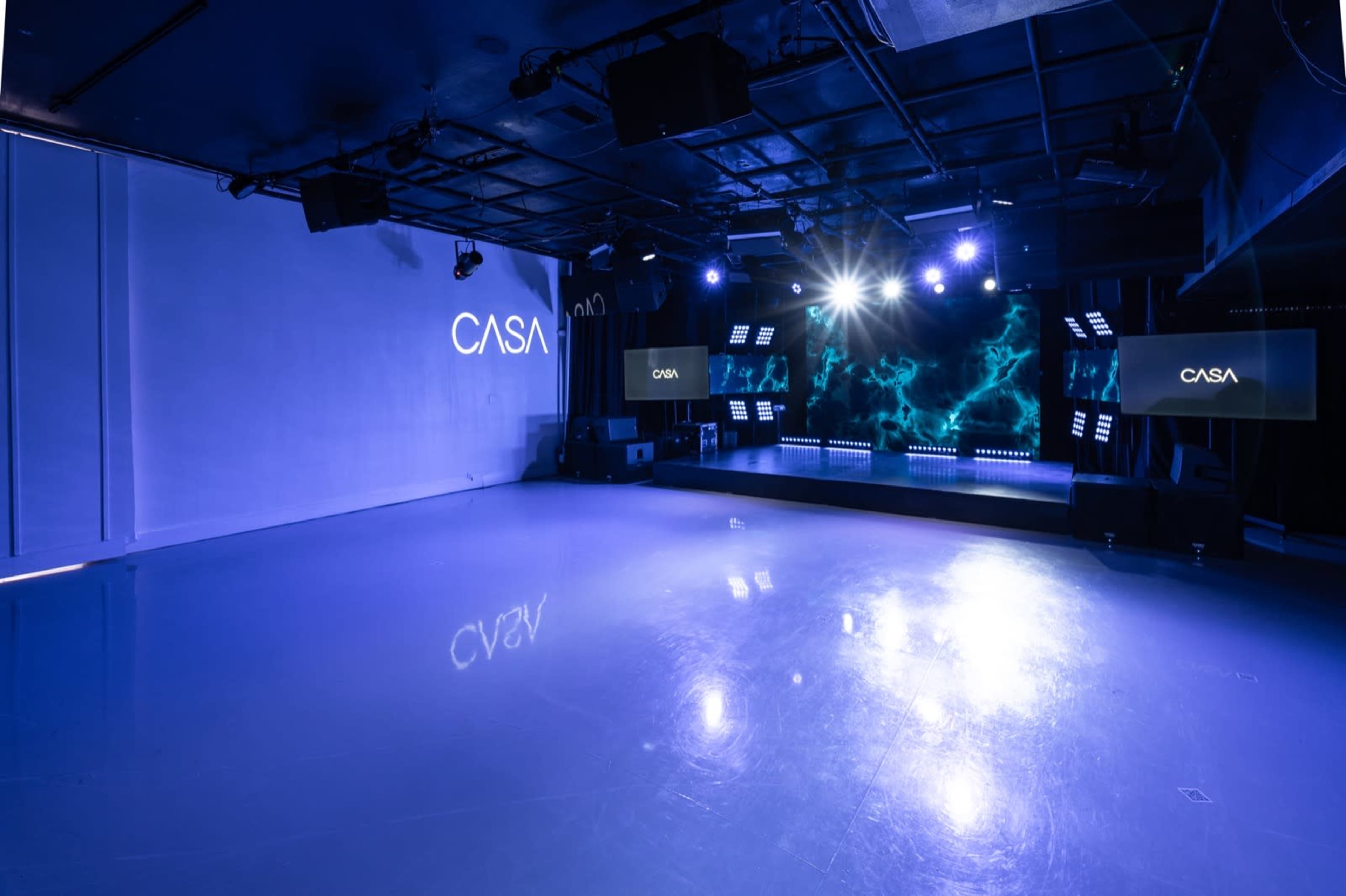 The image shows an empty event space with a polished floor, featuring a stage with lighting and screens on either side, and the word "CASA" displayed prominently.