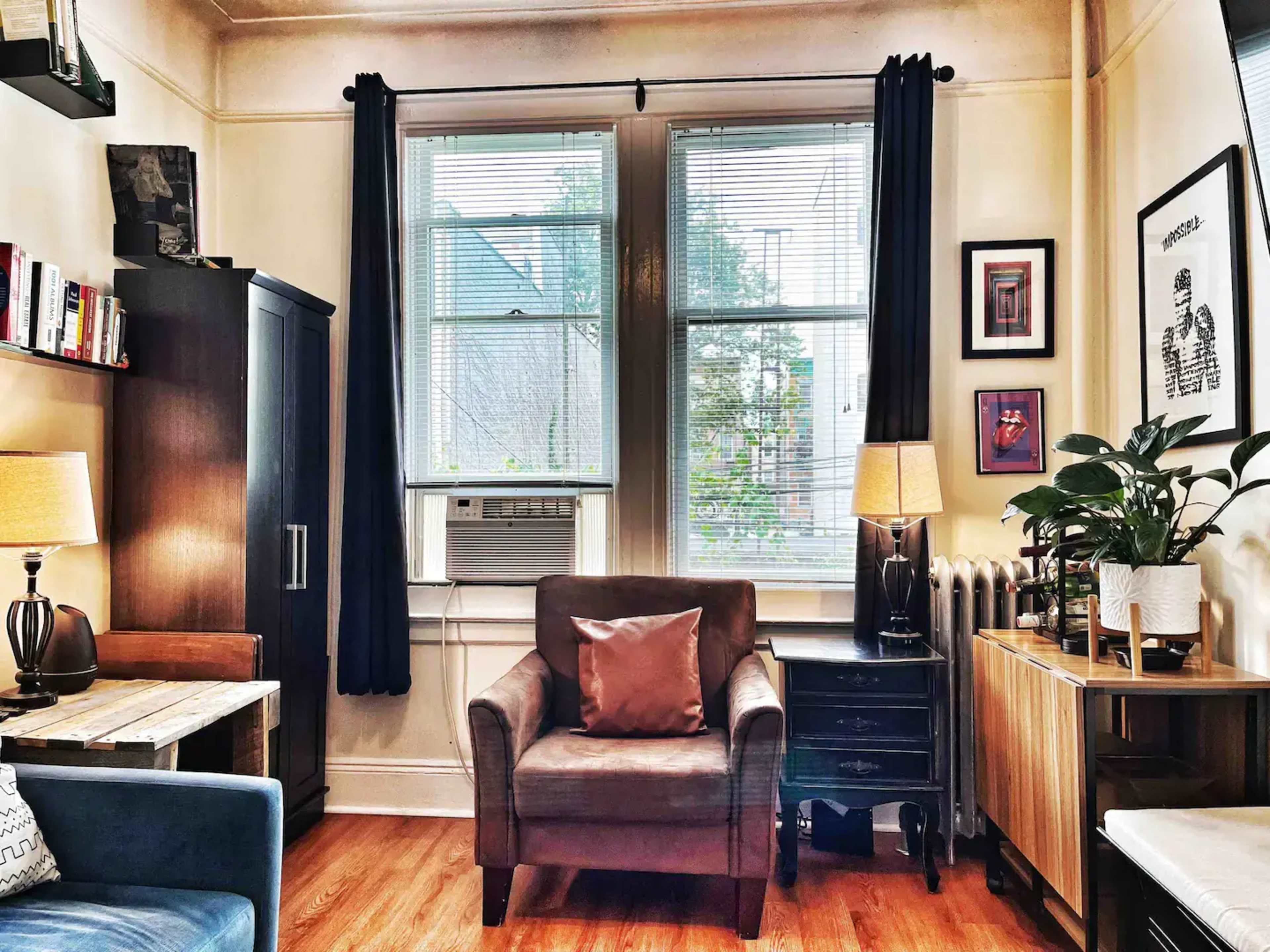 A cozy living room with two windows, an air conditioning unit, a brown armchair, a wooden table, and various decorative elements including plants and lamps.