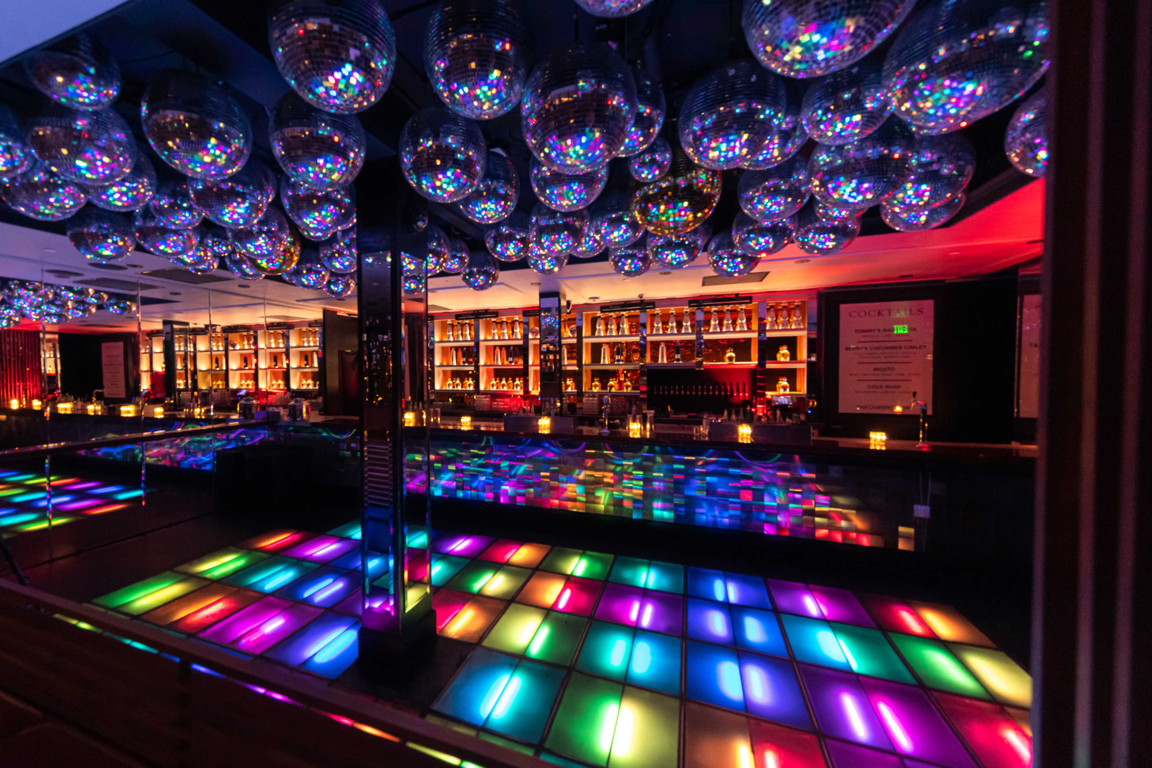 A vibrant dance floor illuminated with colorful lights, surrounded by mirrored disco balls and a well-stocked bar in the background.