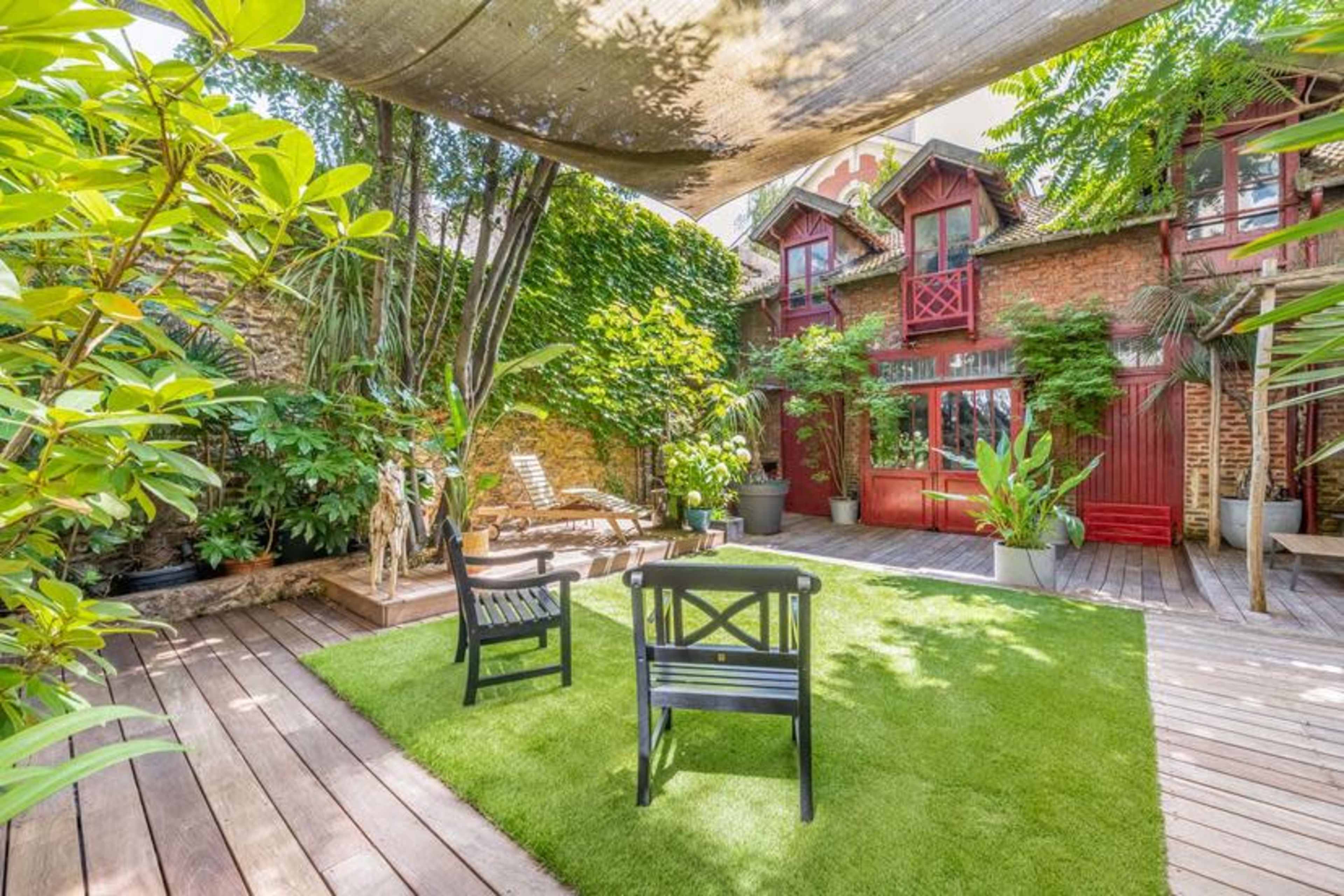 A well-maintained garden area with wooden decking, green grass, and two black chairs surrounded by vibrant plants and a red-brick building in the background.