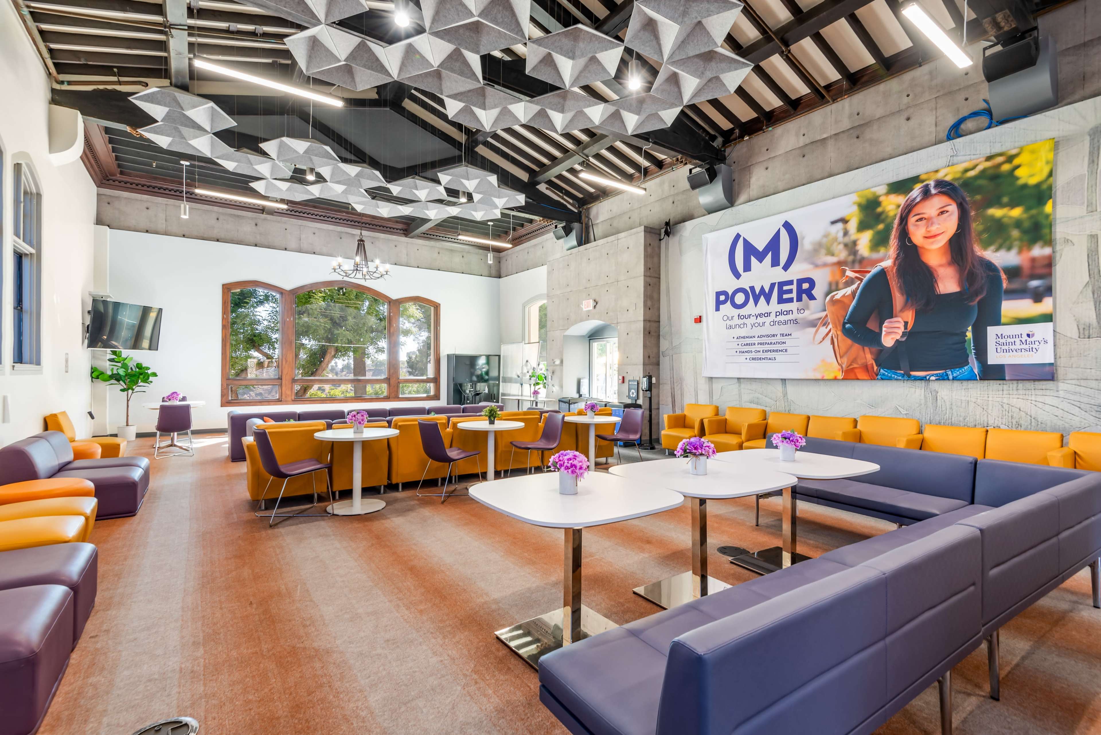 The image shows a modern meeting space with purple and orange seating, large windows, decorative ceiling elements, and a screen displaying a promotional graphic.