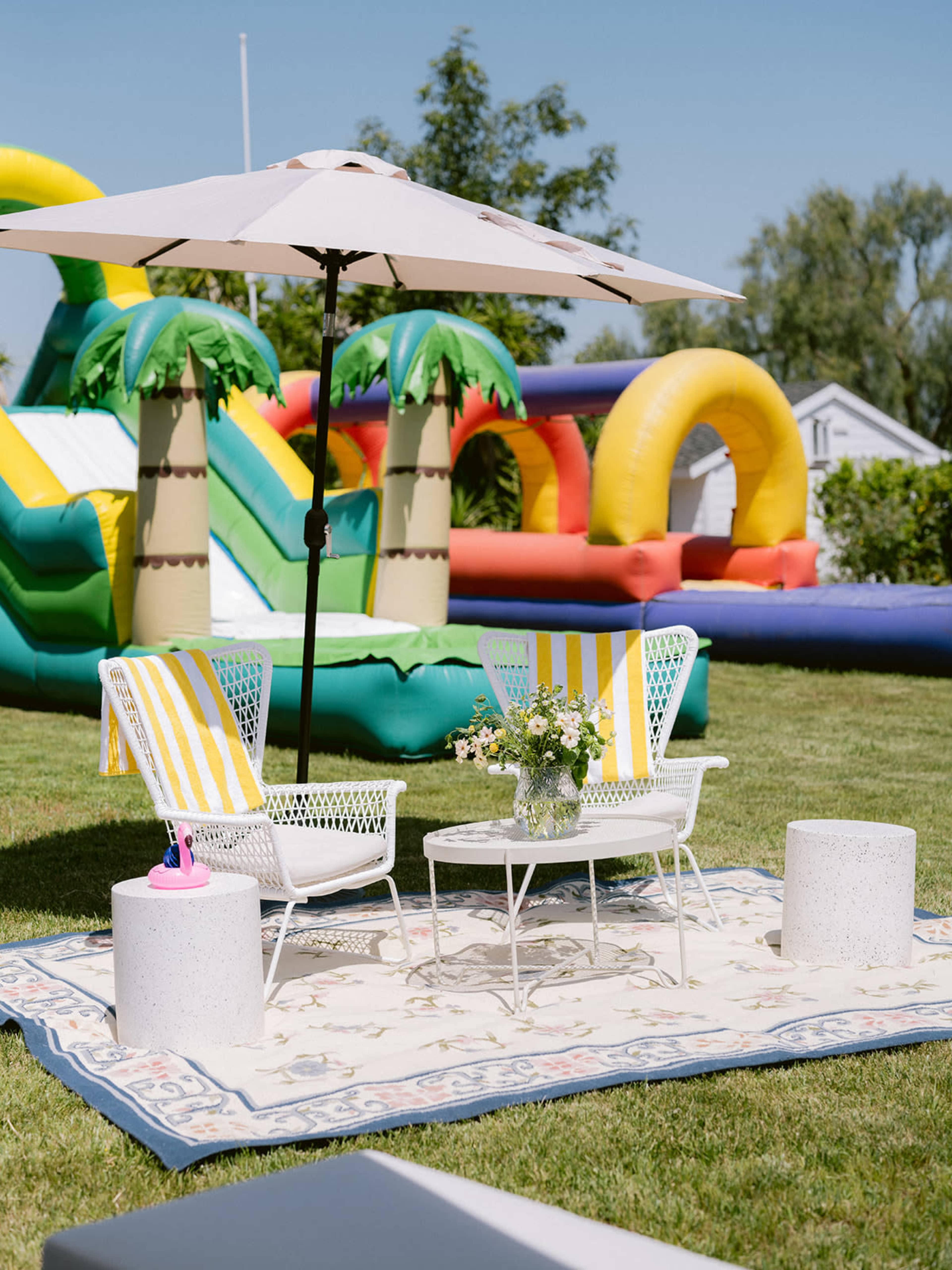 A patio area with two white chairs and a table sits on a decorative rug under an umbrella, surrounded by inflatable structures and greenery.