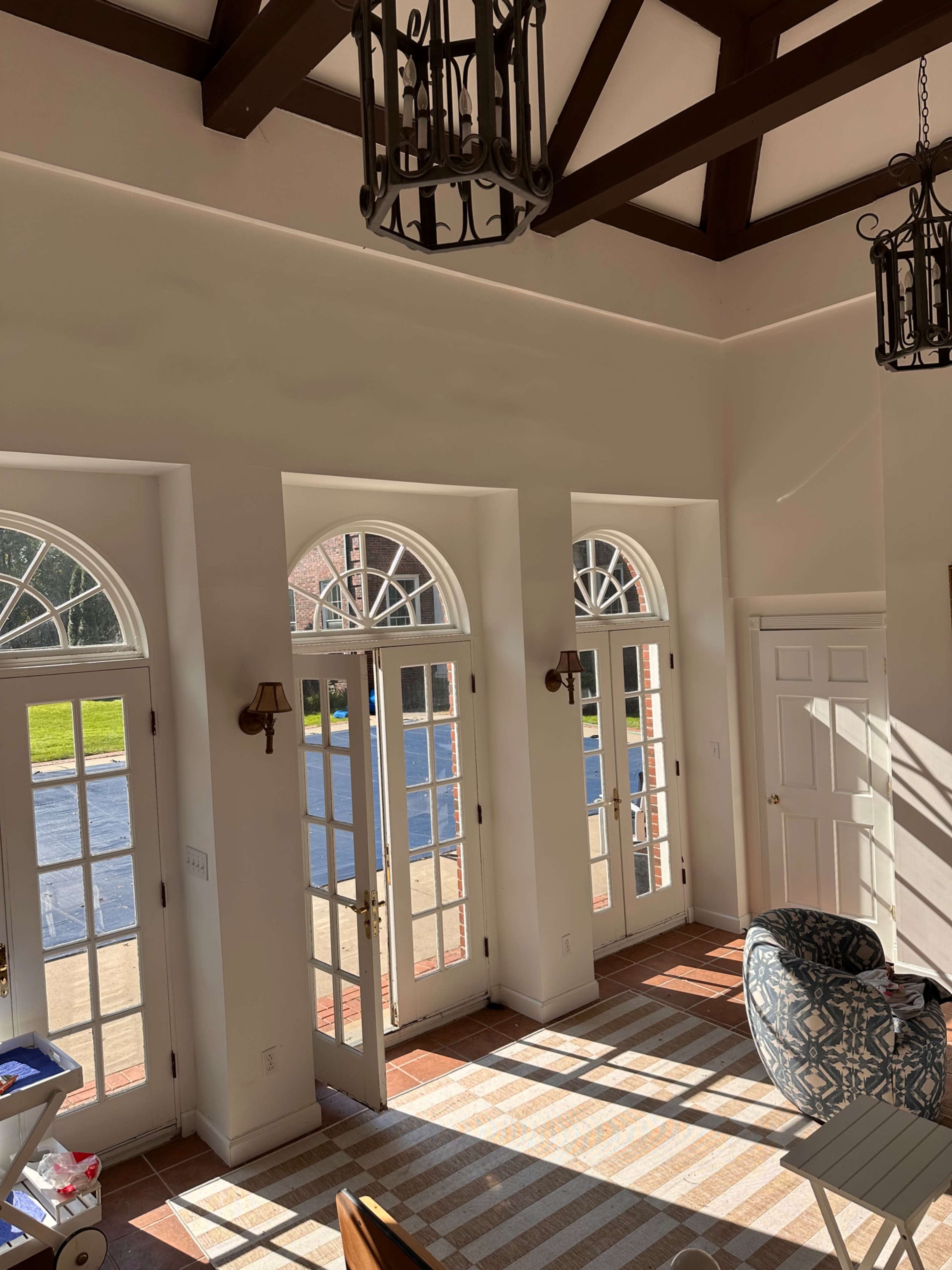 The image shows a brightly lit room with large arched windows, wooden beams on the ceiling, and a striped rug on the floor.