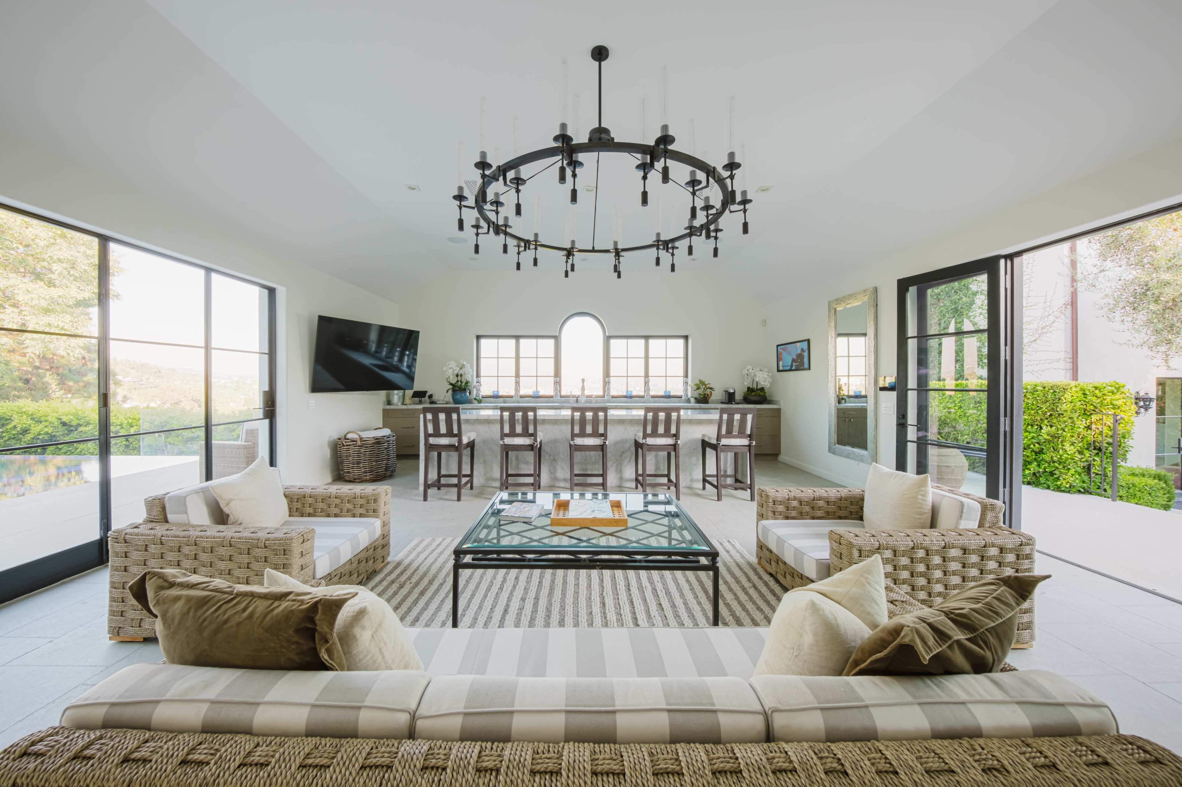 The image depicts a spacious indoor living area with two seating arrangements, a large chandelier, and a bar with high stools in the background, all surrounded by large windows.