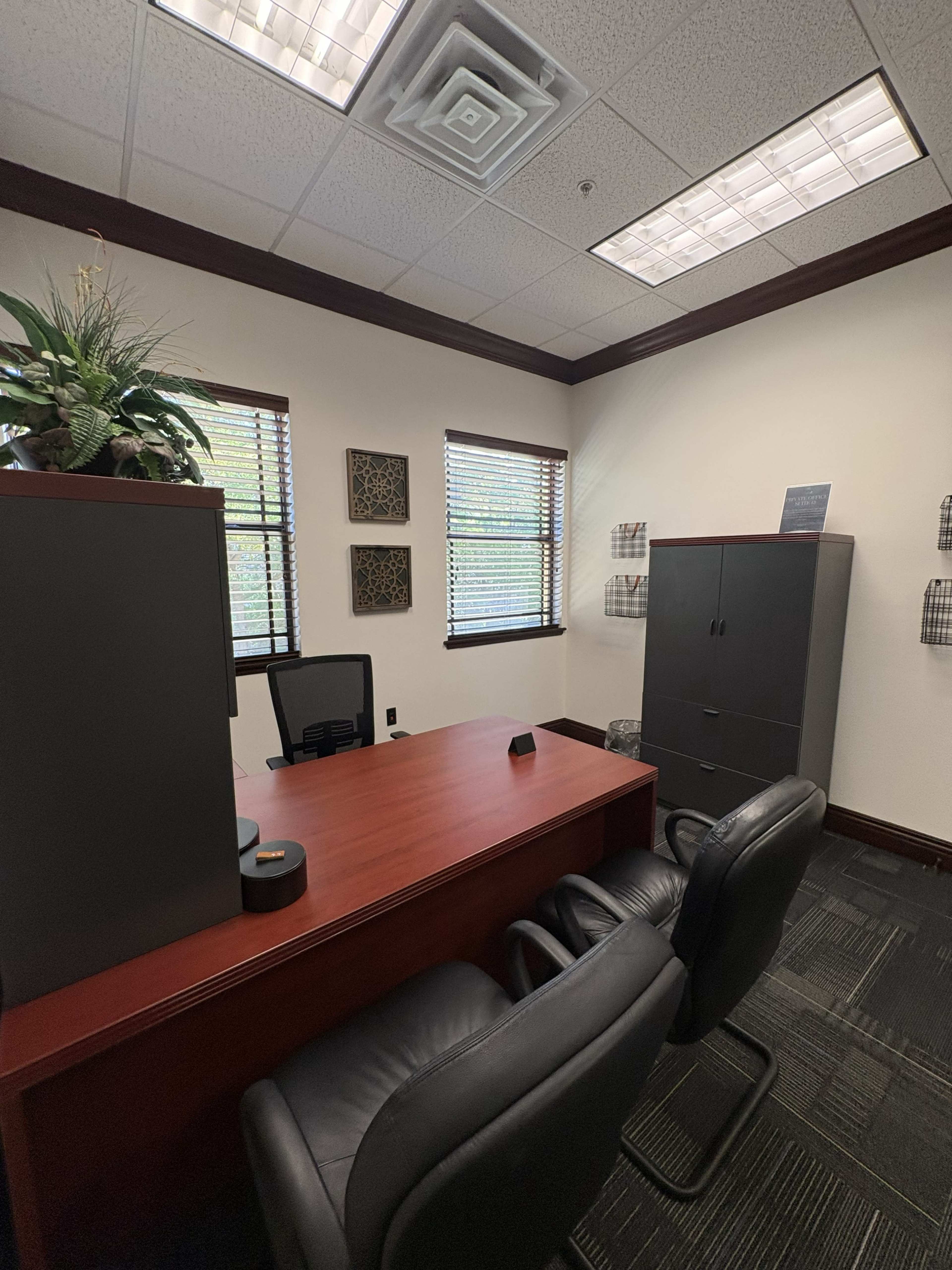 The image shows a small office space with a wooden desk, two black chairs, a filing cabinet, and decorative wall art near the windows.