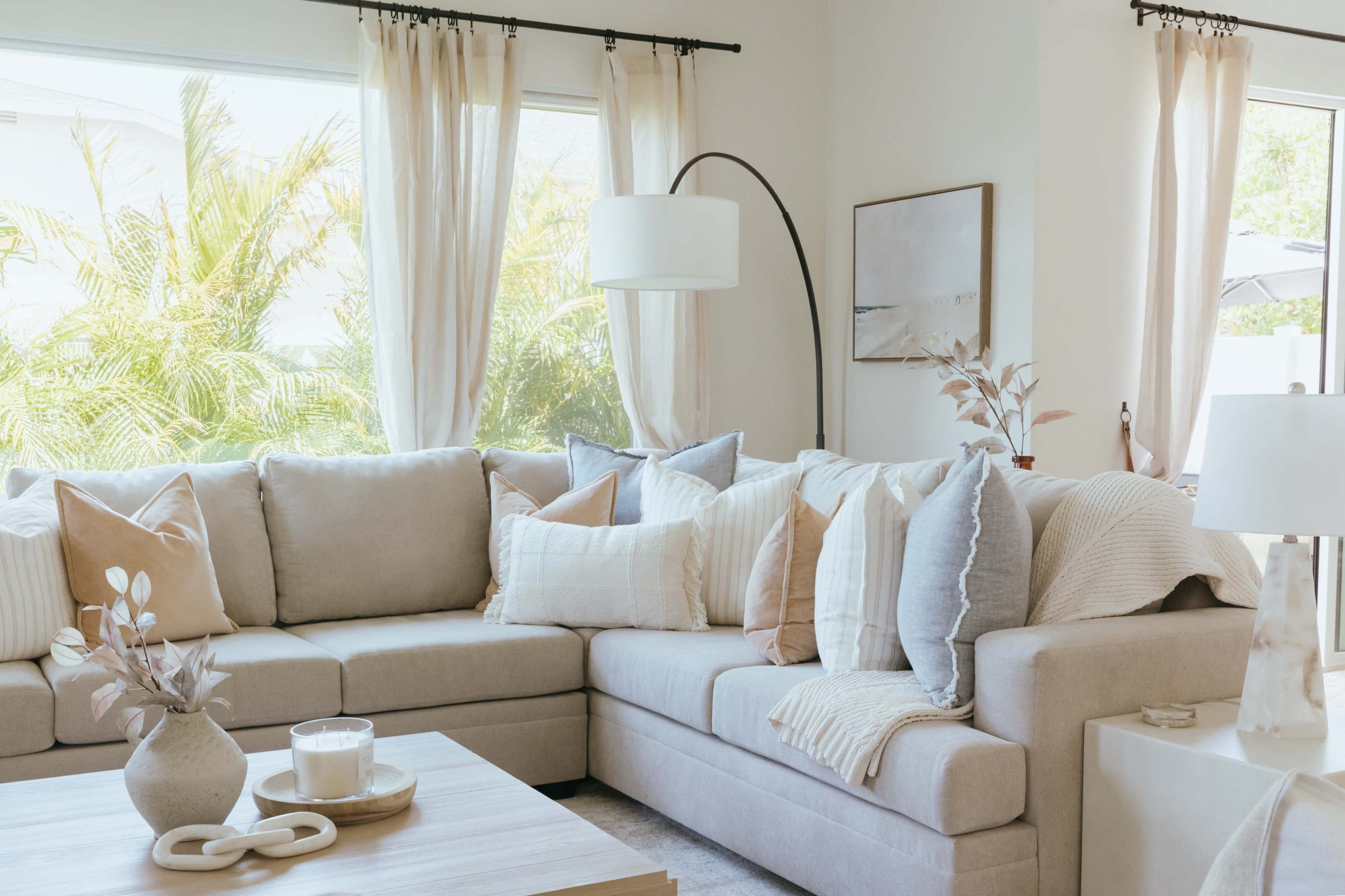 A light-filled living room features a large, beige sectional sofa with multiple decorative pillows and a coffee table adorned with a candle and a vase.