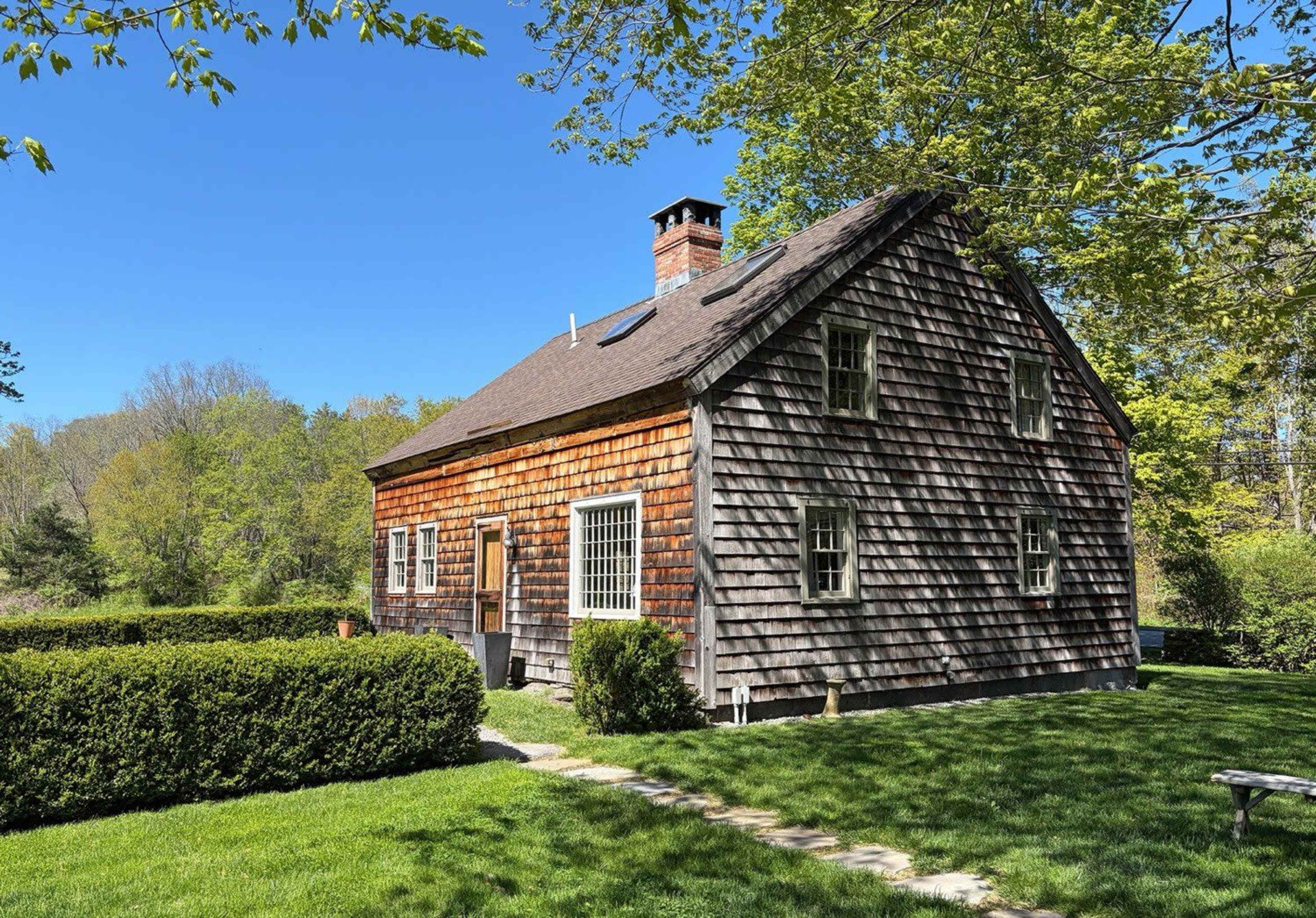 A weathered wooden house with a sloped roof and a chimney is surrounded by neatly trimmed hedges and a grassy yard under a clear blue sky.