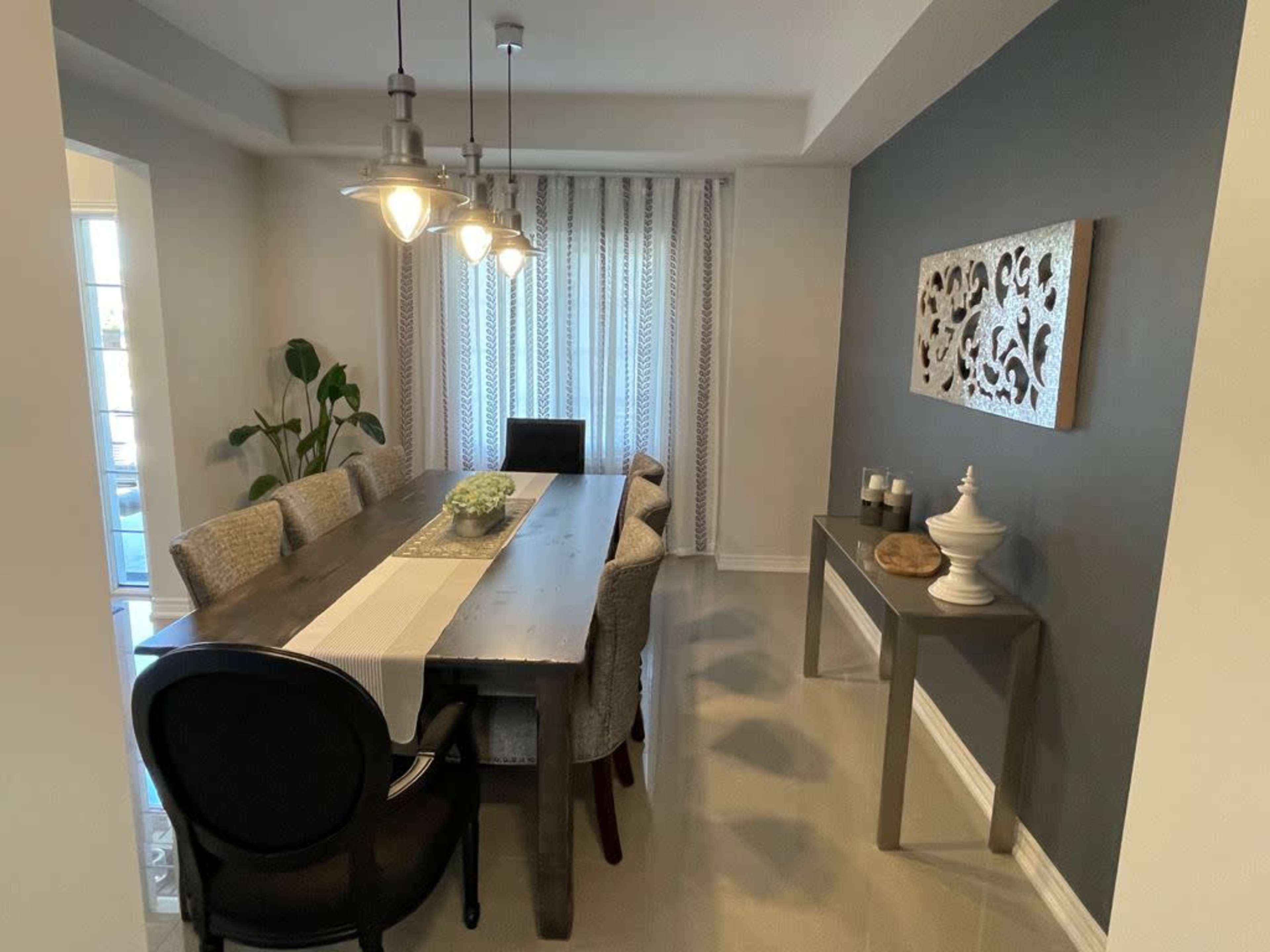A dining area features a long wooden table with upholstered chairs, pendant lighting, and decorative wall art.