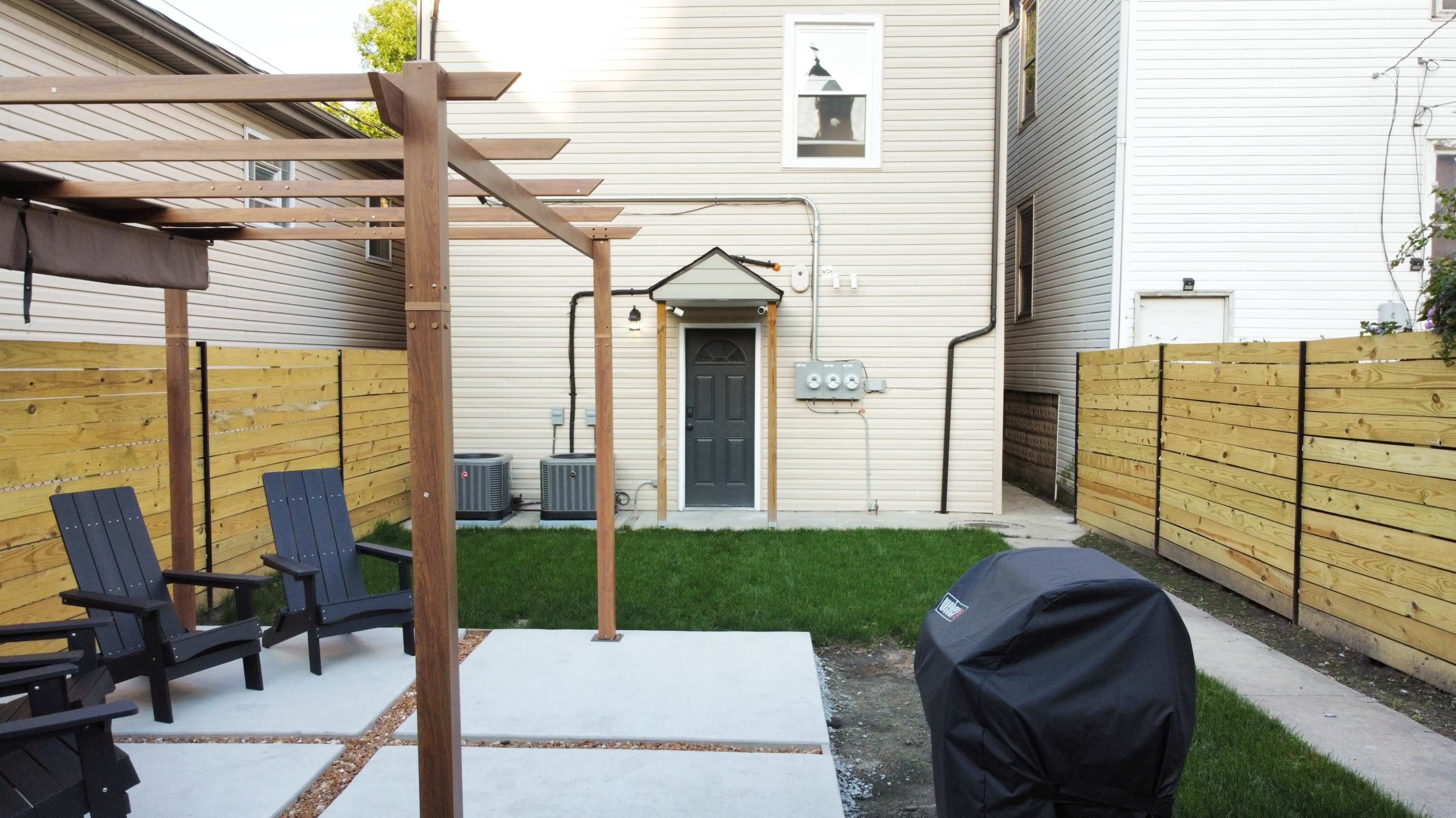 A backyard with a pergola, two black chairs, a barbecue grill, and a neatly maintained patch of grass adjacent to a house.