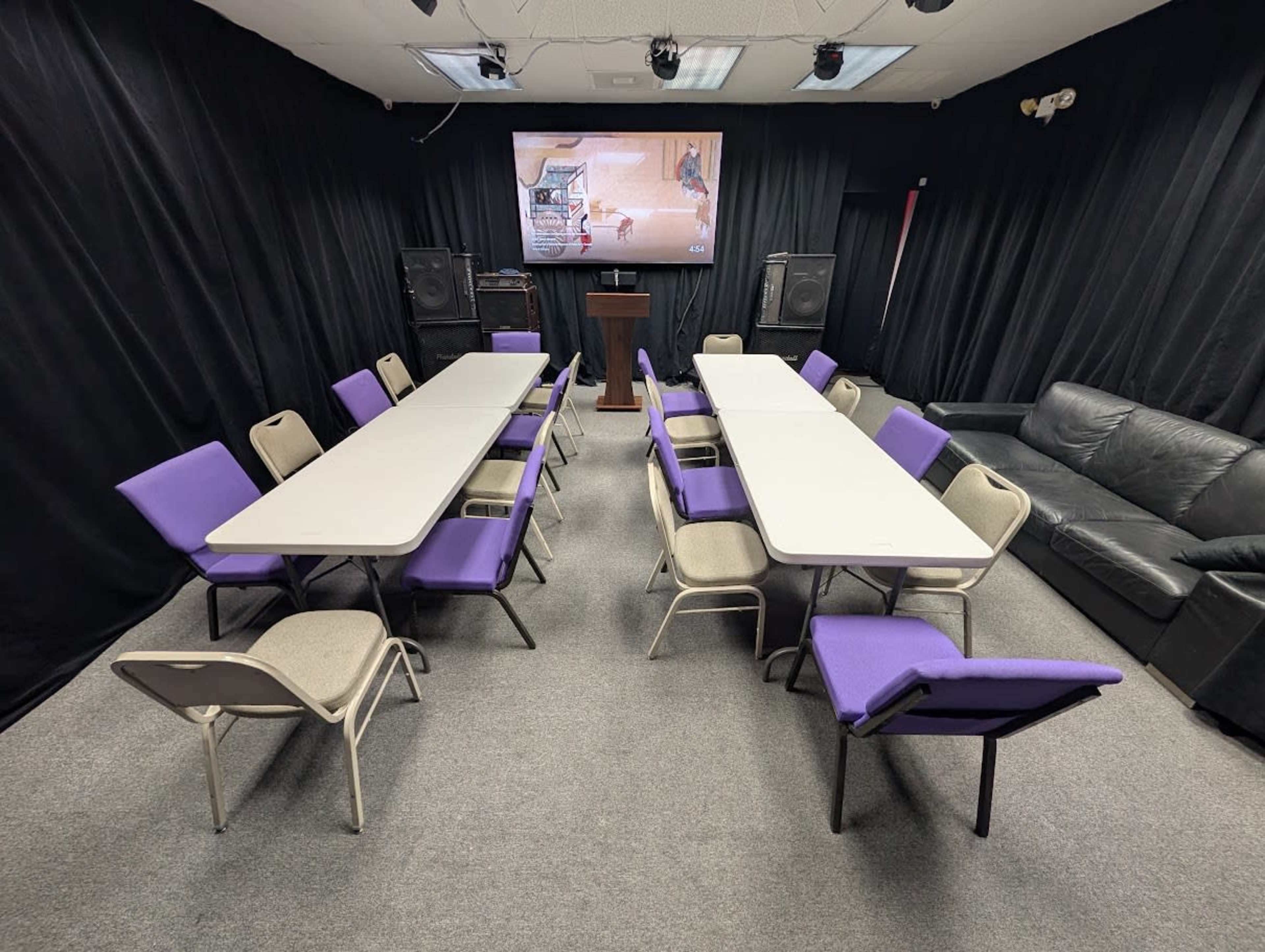 A conference room is set up with several long tables arranged in a U-shape, surrounded by chairs, with a black curtain backdrop and a screen displaying a video.