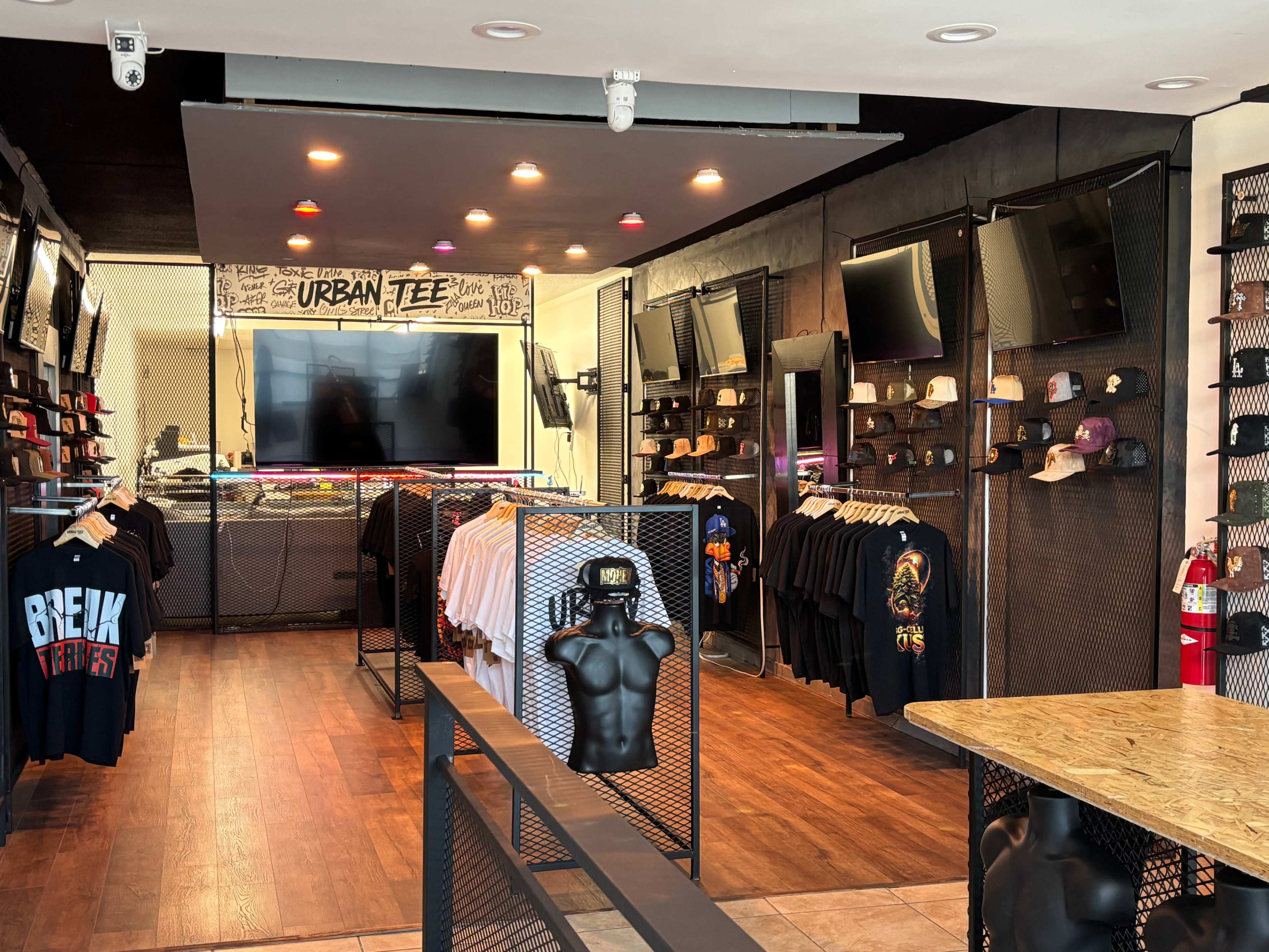 The image shows a retail store interior with clothing racks displaying T-shirts and hats, and multiple screens mounted on the walls.