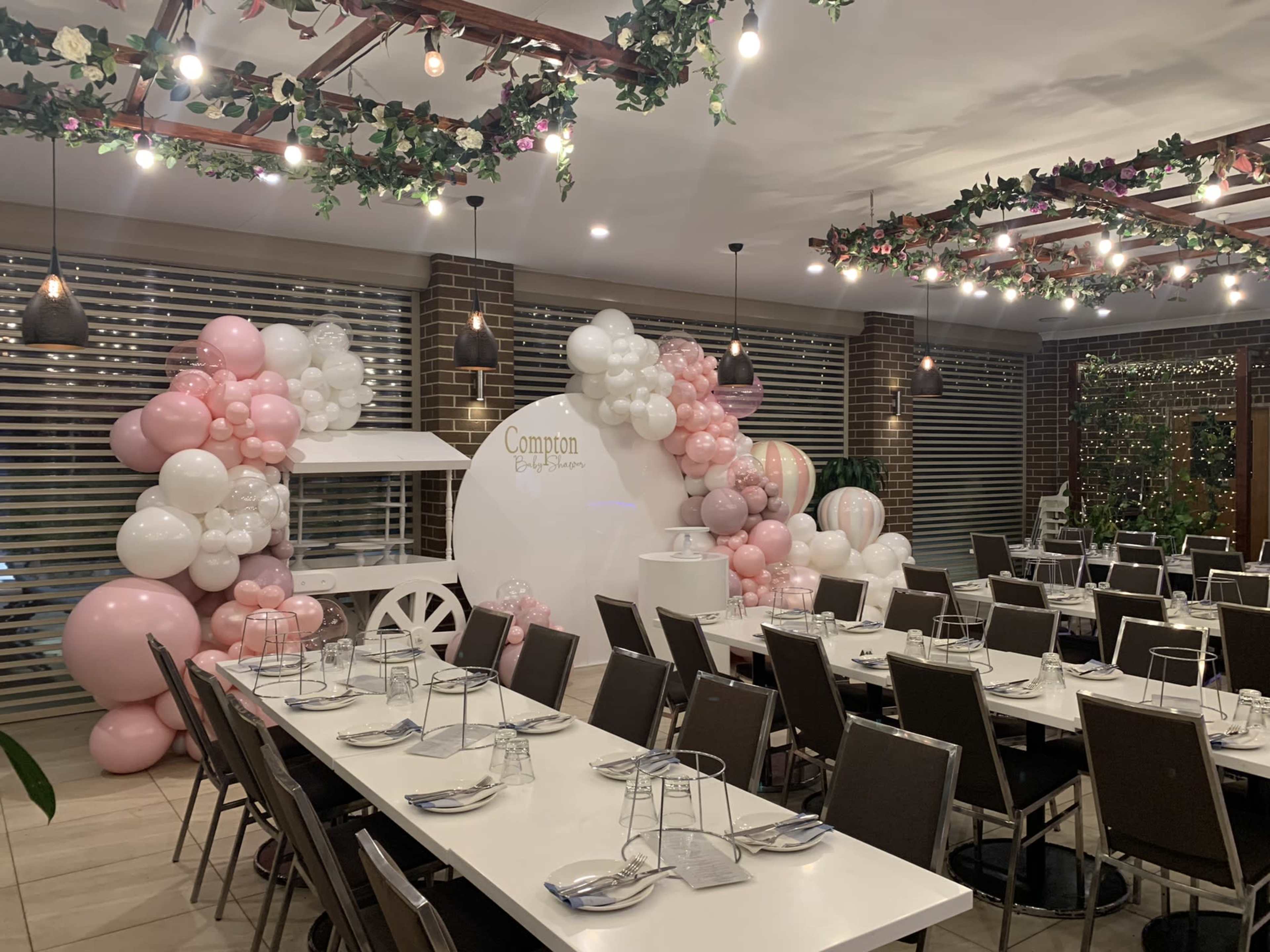 A banquet room is decorated with balloons in pink and white, featuring a carriage display and set with long tables for dining.