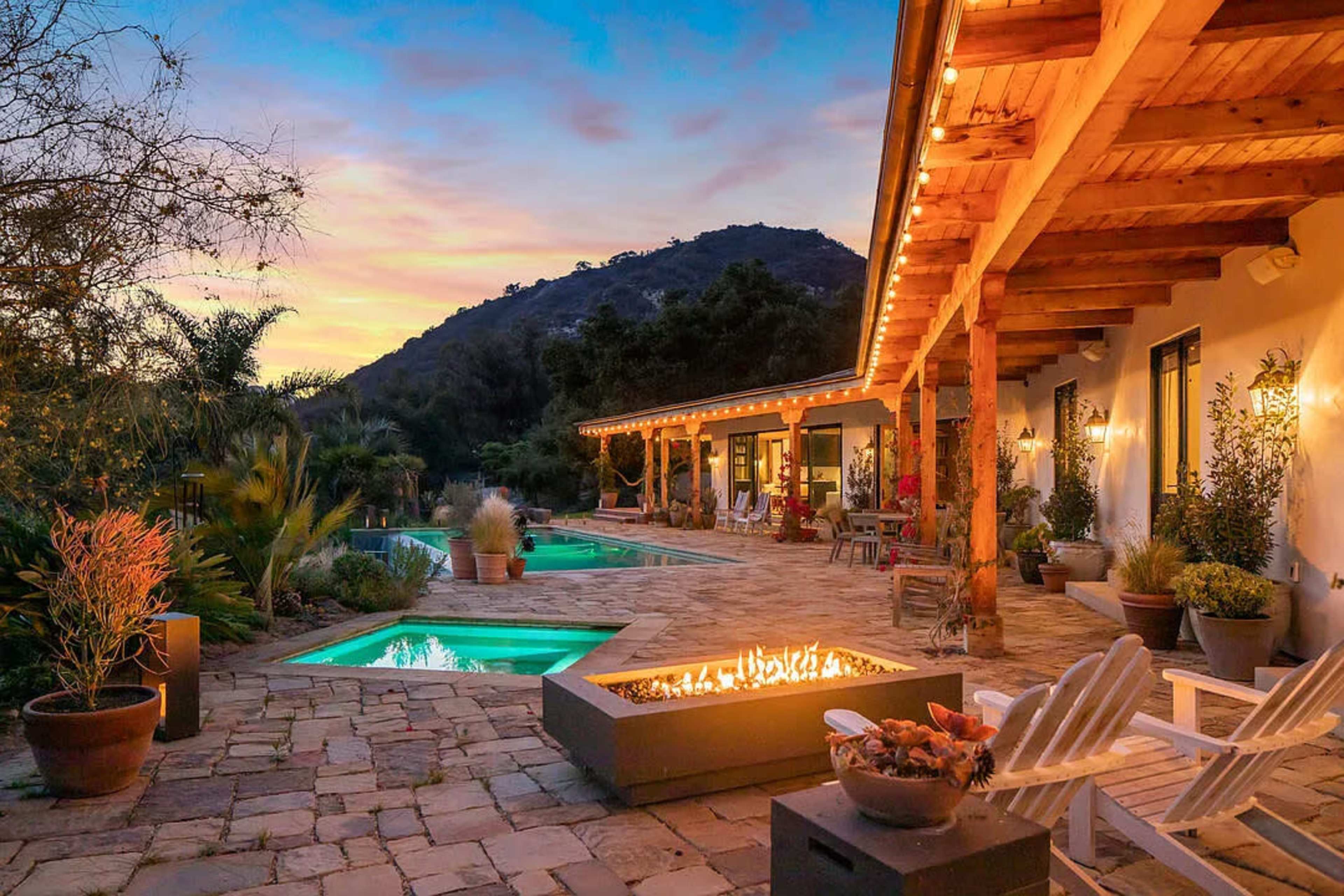 The image shows a scenic outdoor patio area with two swimming pools, a fire pit, and a mountain backdrop during sunset.
