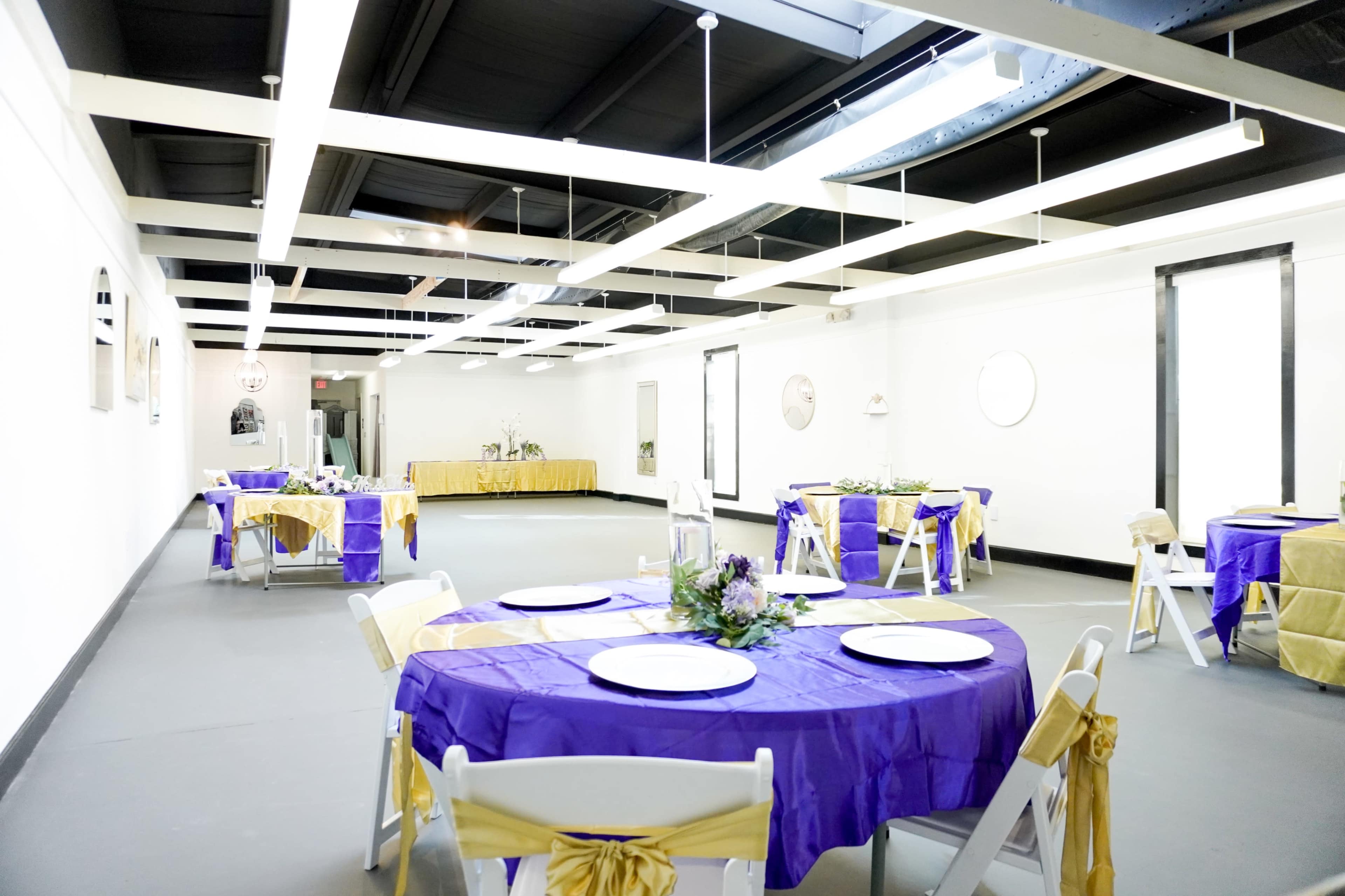 The image shows a spacious event venue with purple and gold table settings, including round tables covered with tablecloths and decorated with floral arrangements.