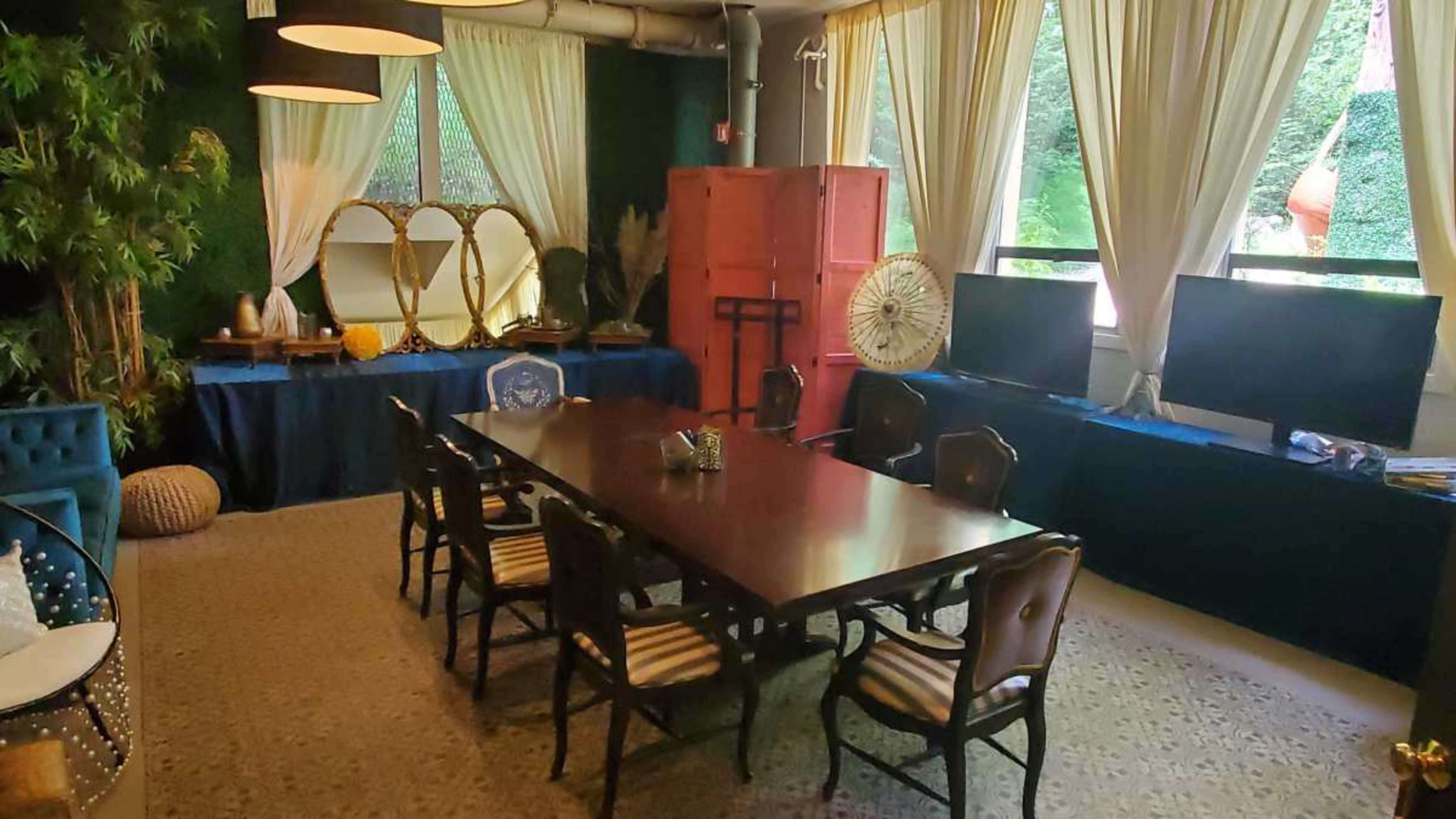 A room furnished with a large wooden table surrounded by chairs, two televisions on a blue-covered surface, and decorative elements including plants and curtains.