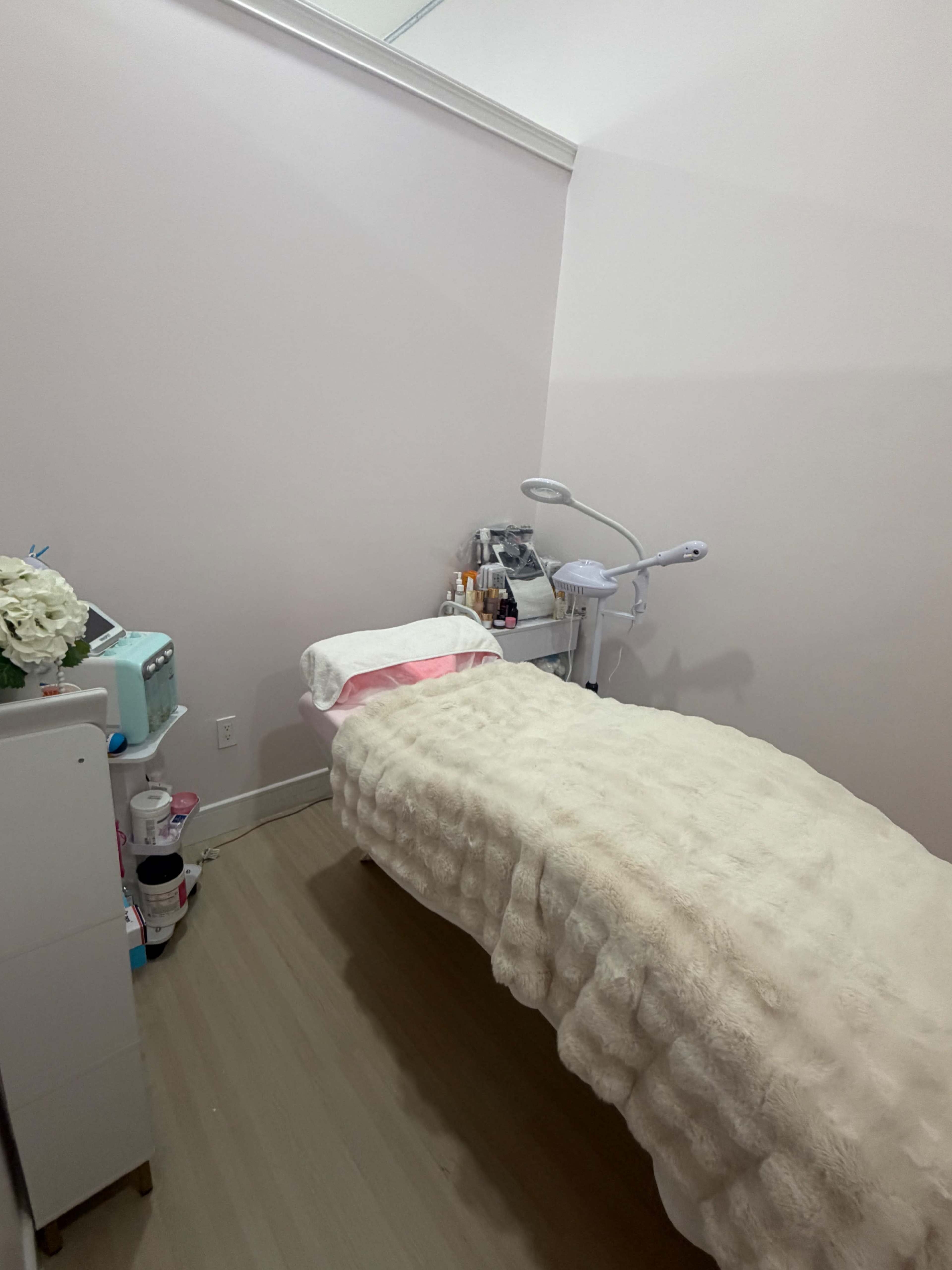 A small room is set up with a treatment bed covered in a soft, light-colored blanket, alongside a table with various cosmetic products and a lamp.