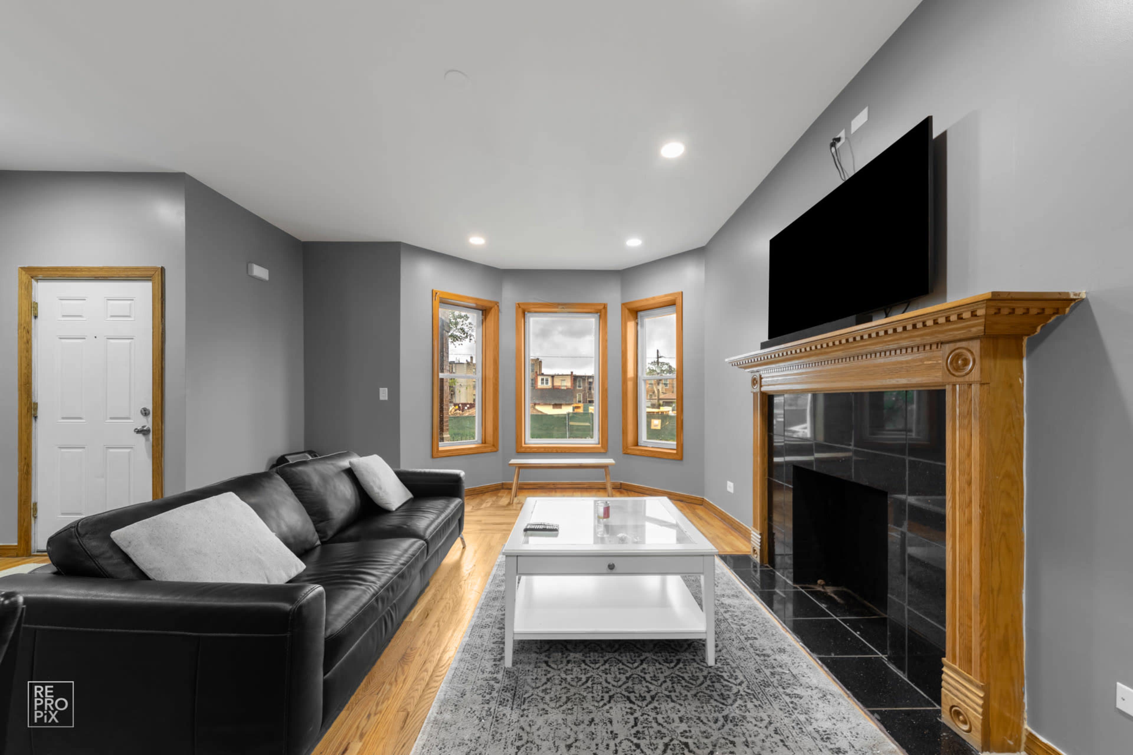 The living room features a black leather sofa, a white coffee table, and three large windows set in a gray wall with a fireplace underneath a mounted TV.