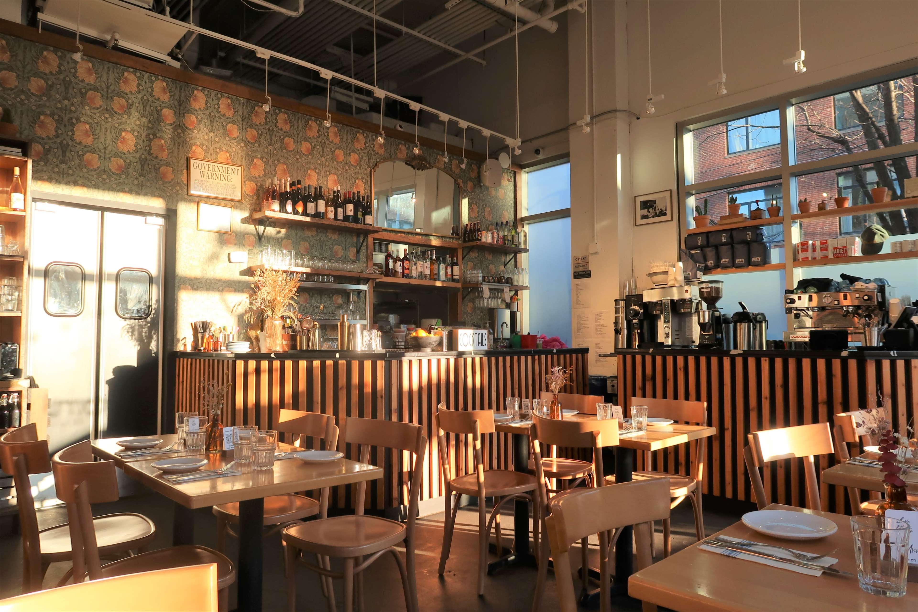 A brightly lit café features wooden tables and chairs, a bar with coffee machines, and decor adorned with floral wallpaper.