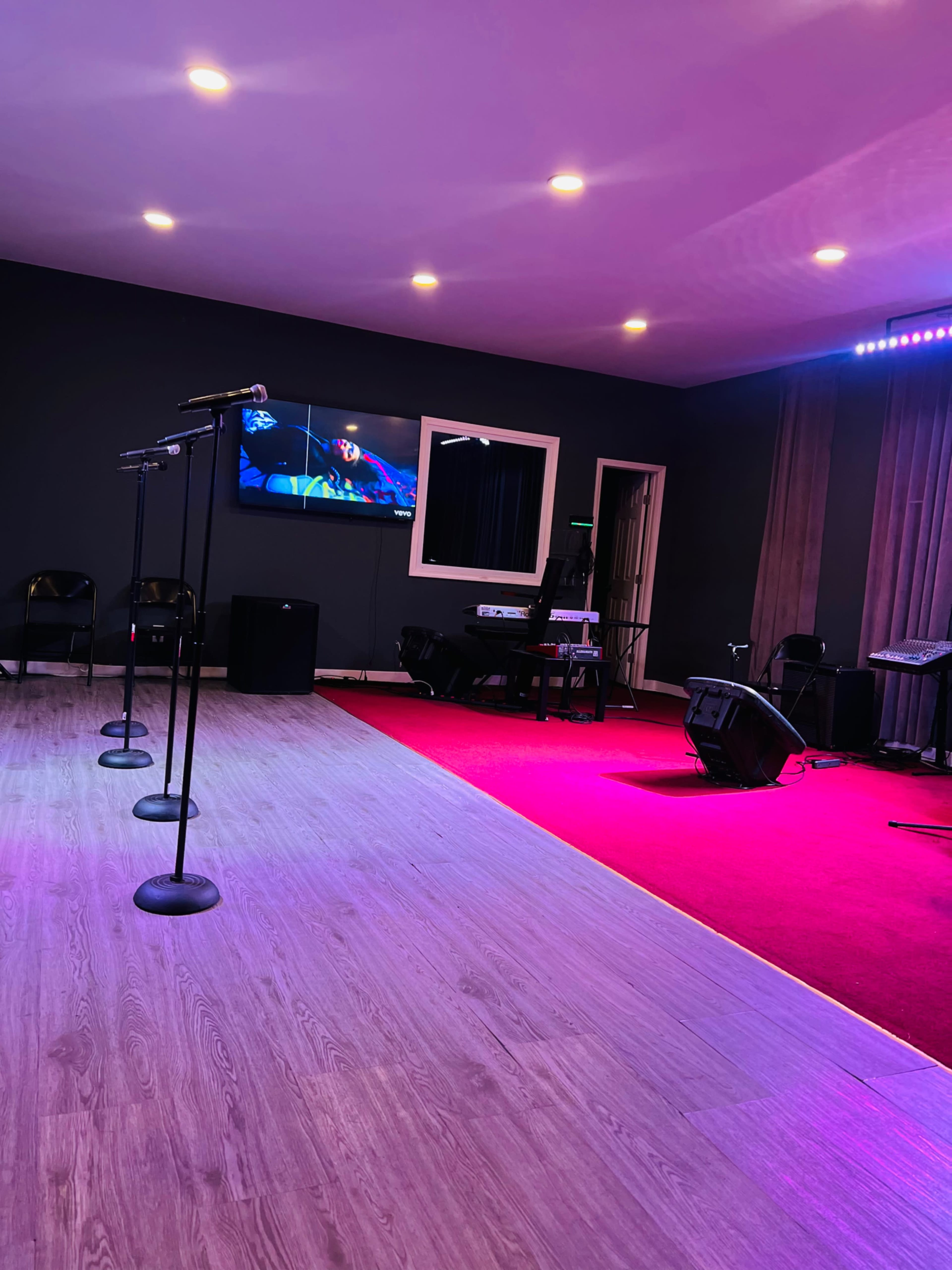 The image shows a small performance space with microphone stands, a television screen, and various musical equipment against a backdrop of contrasting flooring and lighting.