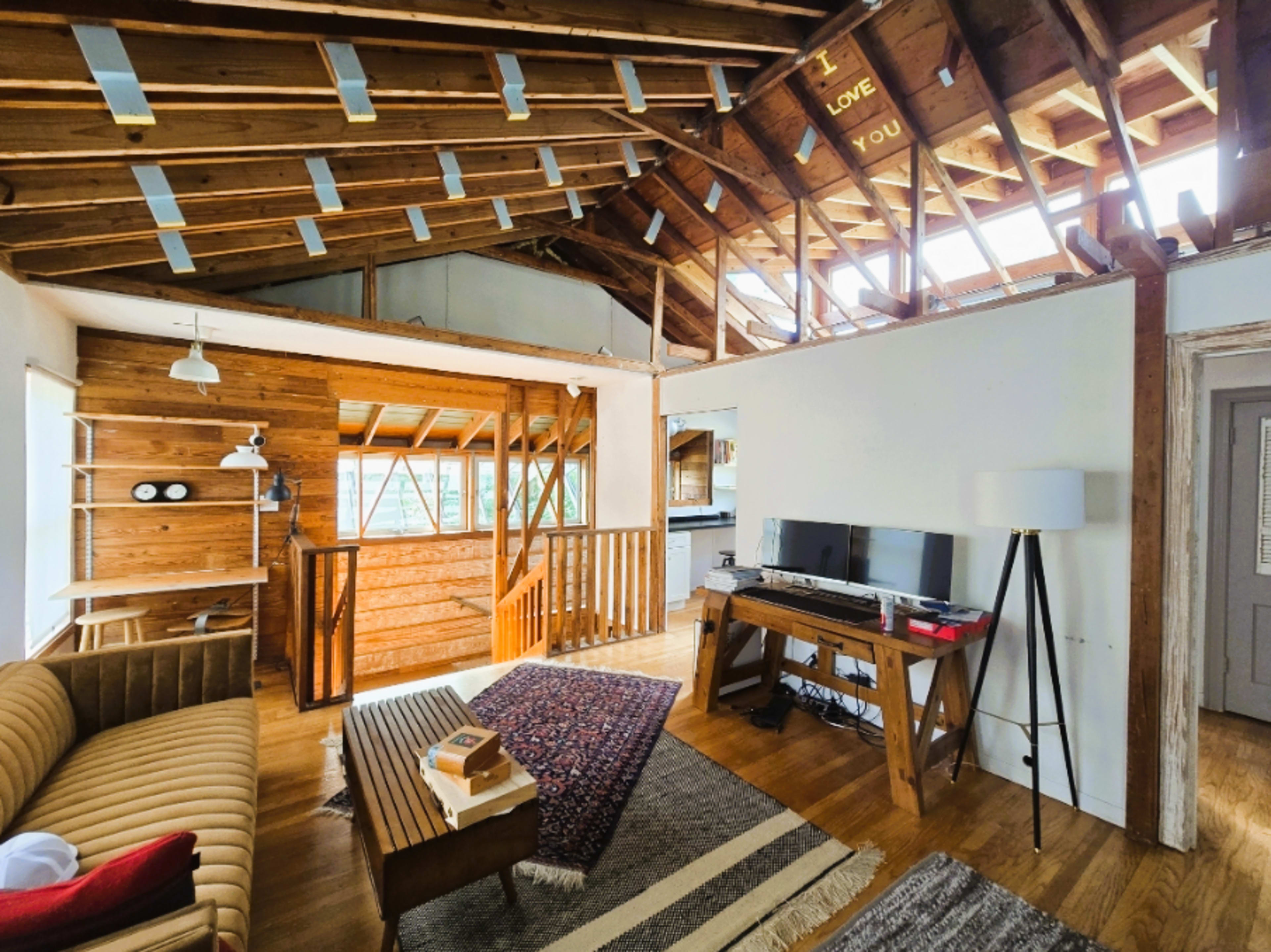 The image shows a rustic living room with wooden beams, a sofa, a desk setup with a computer, and large windows allowing natural light to enter.