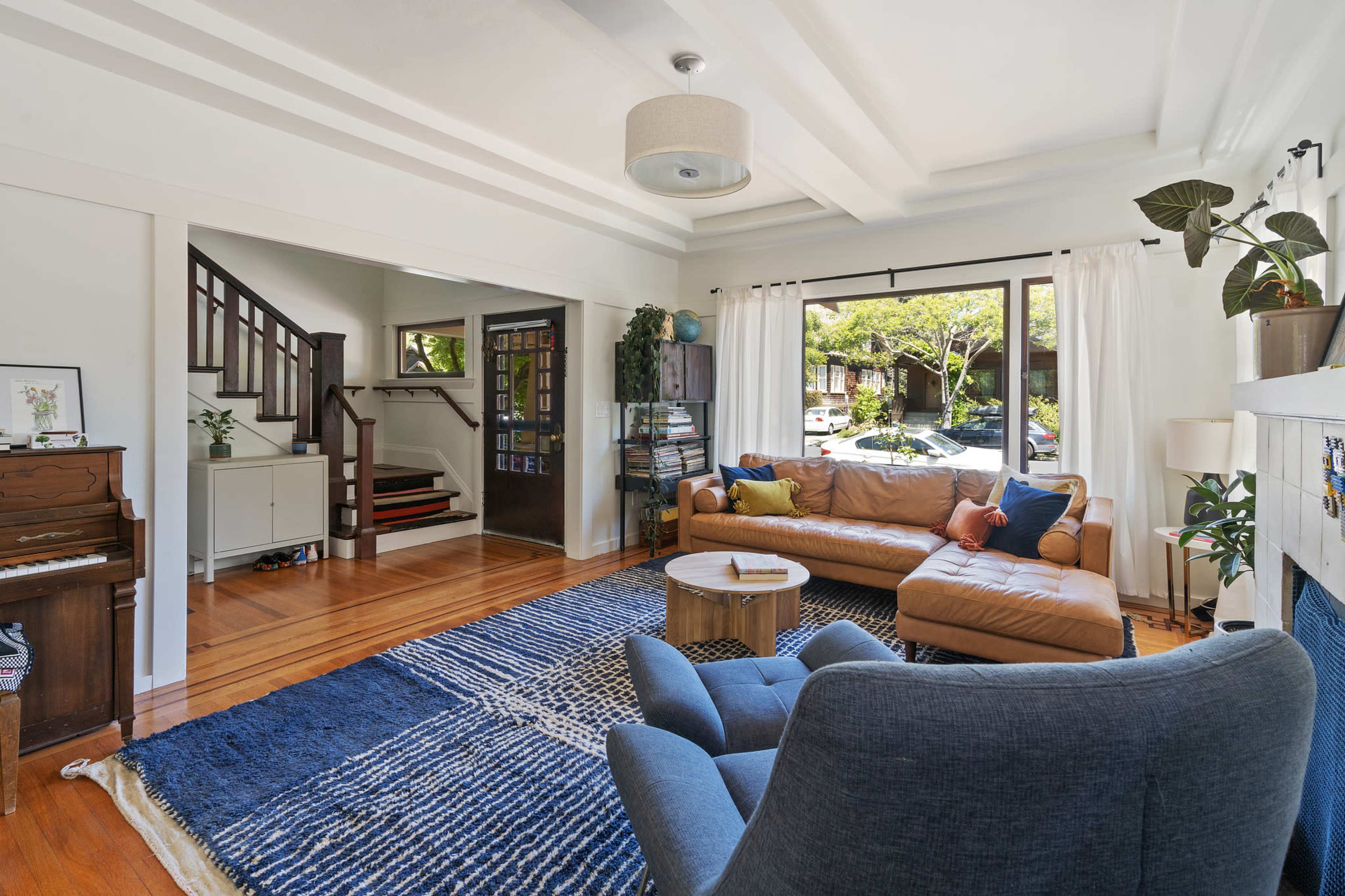 A spacious living room with a leather sofa, a blue armchair, a piano, and large windows overlooking a garden.