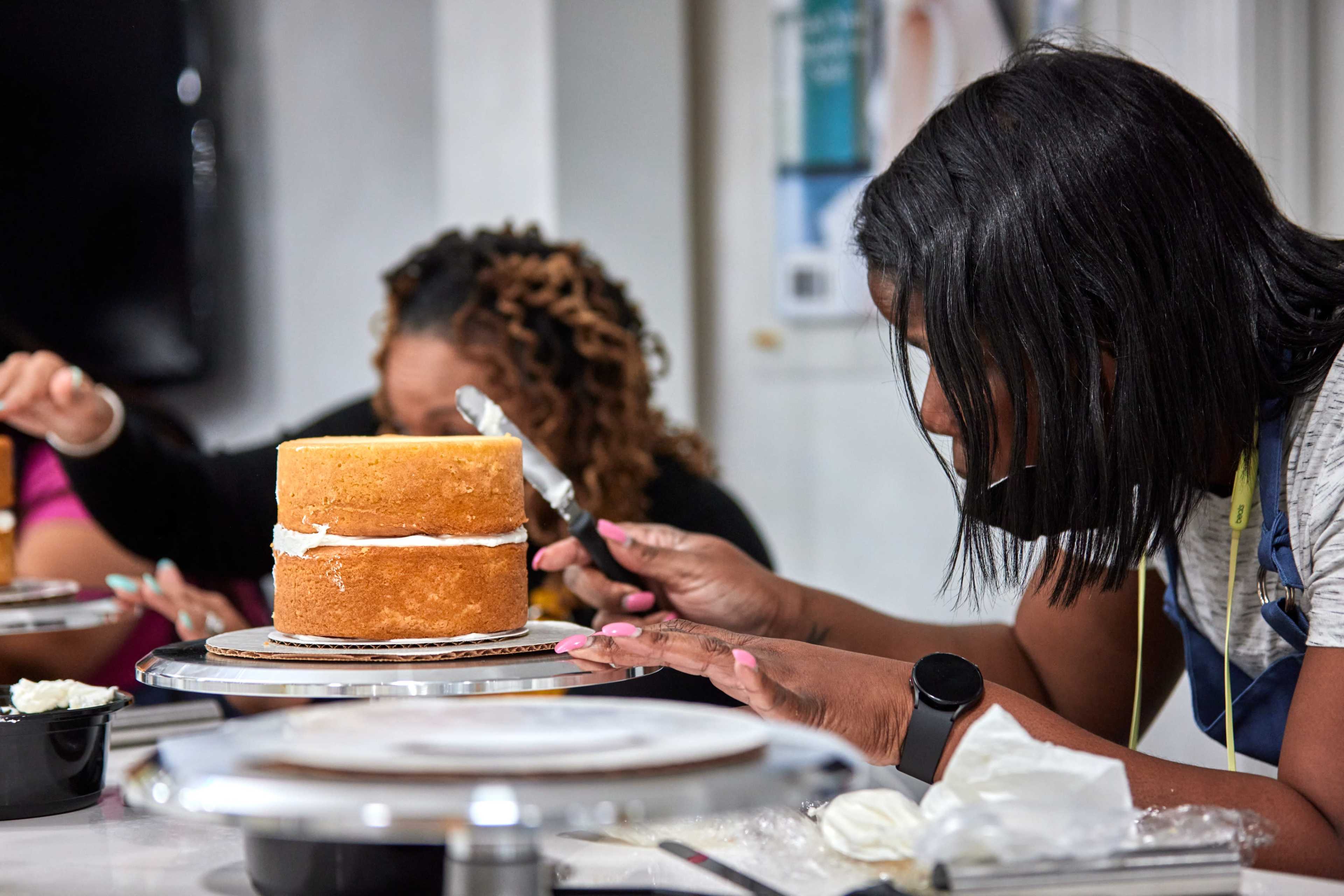 Two individuals are decorating a layered cake with frosting in a brightly lit kitchen setting.