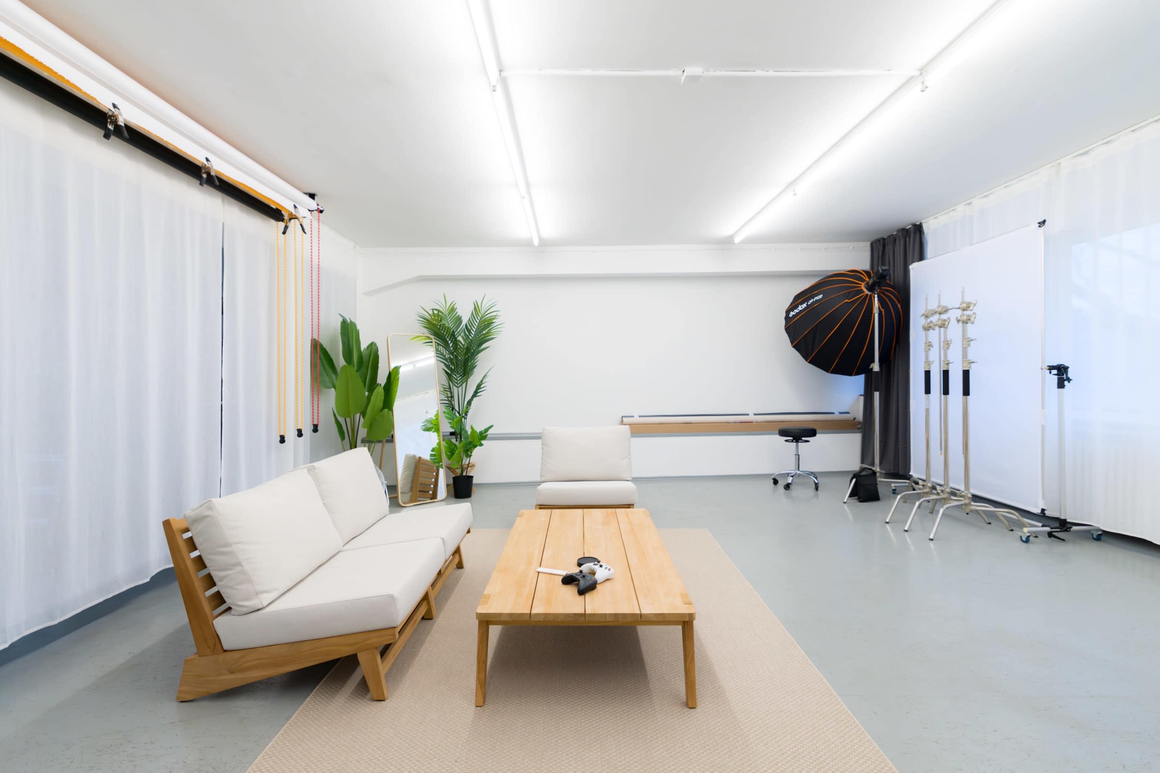The image shows a brightly lit studio interior with a minimalist design, featuring a sofa and coffee table, plants, and photography equipment.