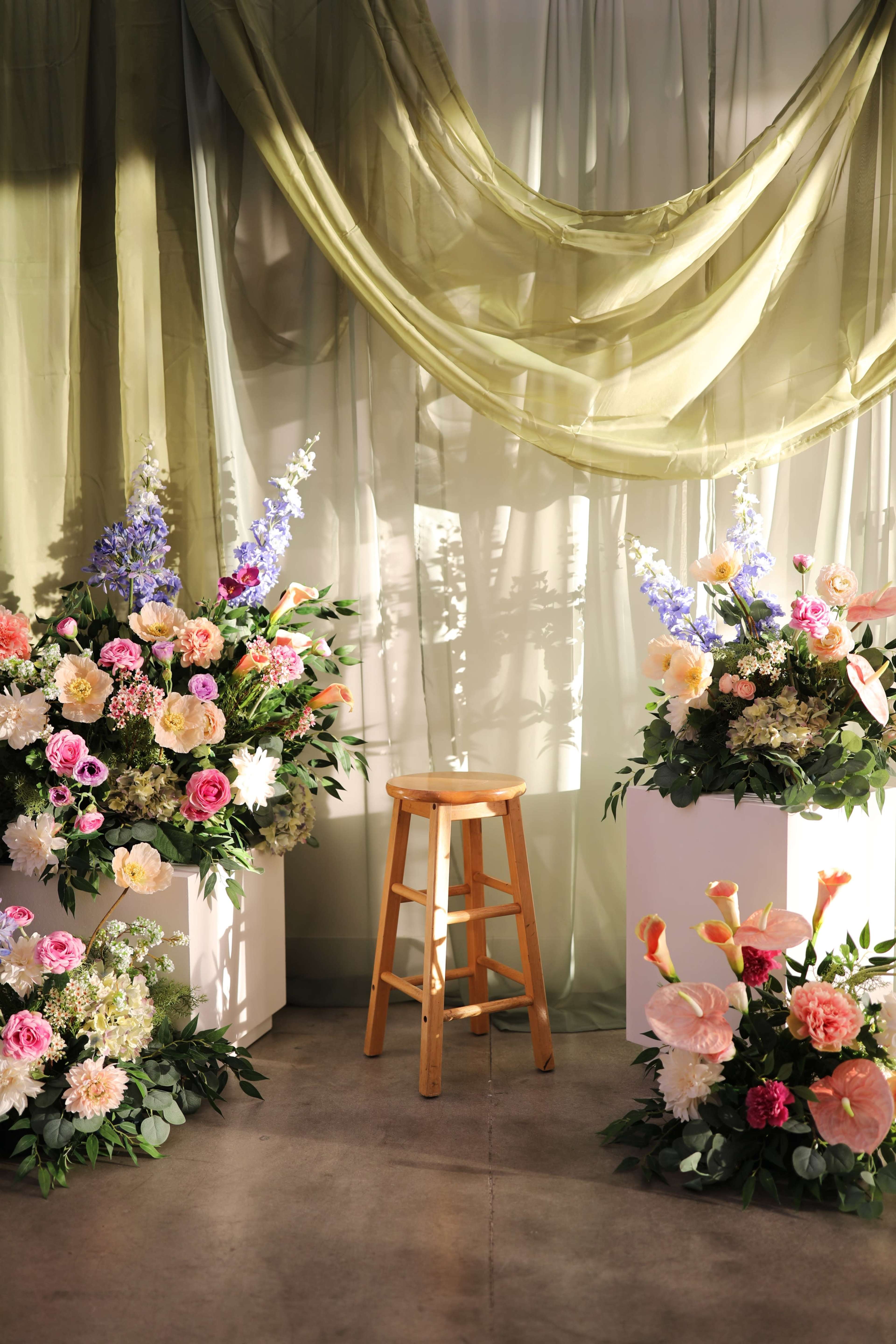 A wooden stool is placed in front of two large flower arrangements surrounded by draped fabric in soft green tones.
