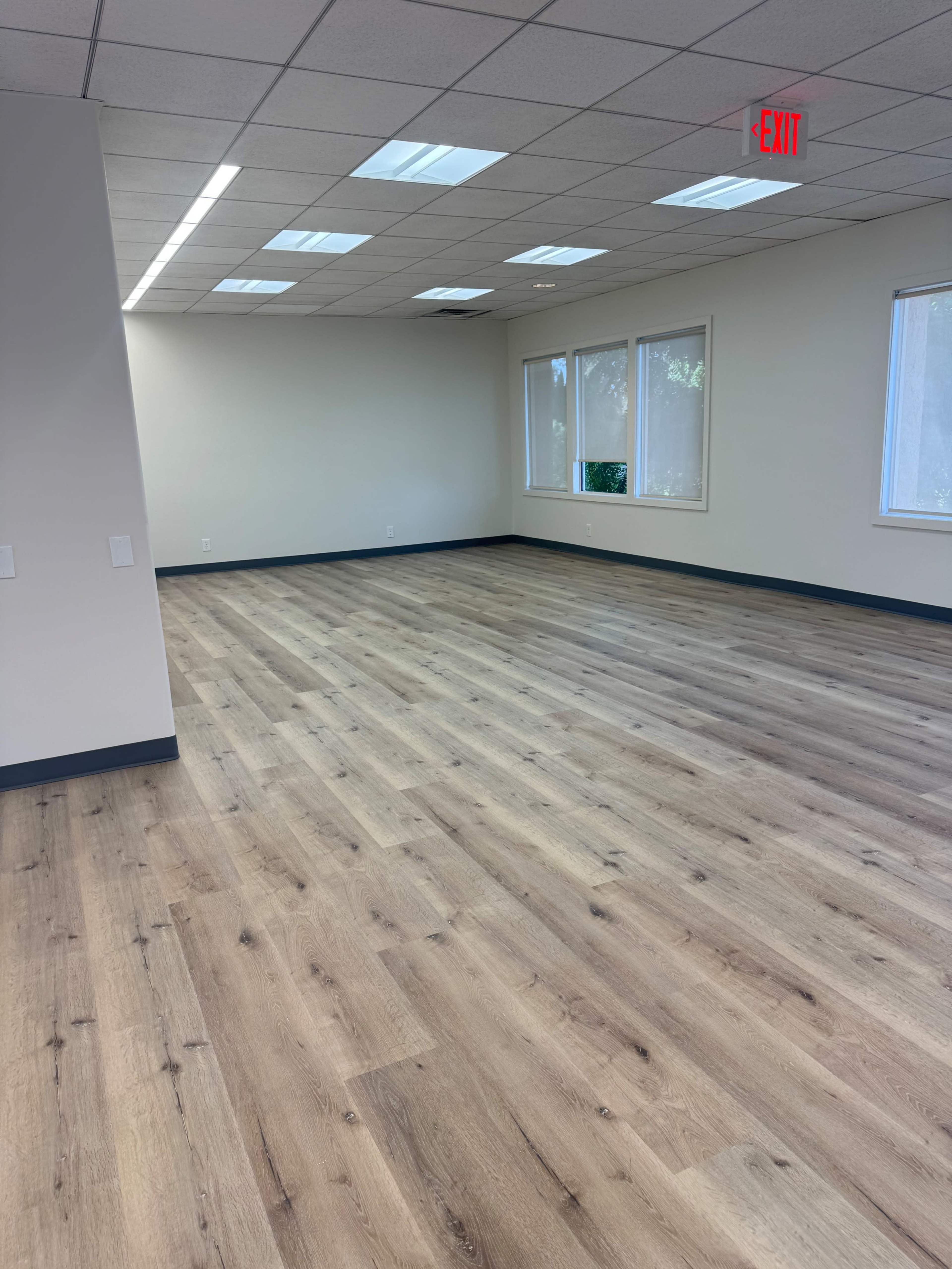 The image shows an empty room with wooden flooring, white walls, and fluorescent ceiling lights.
