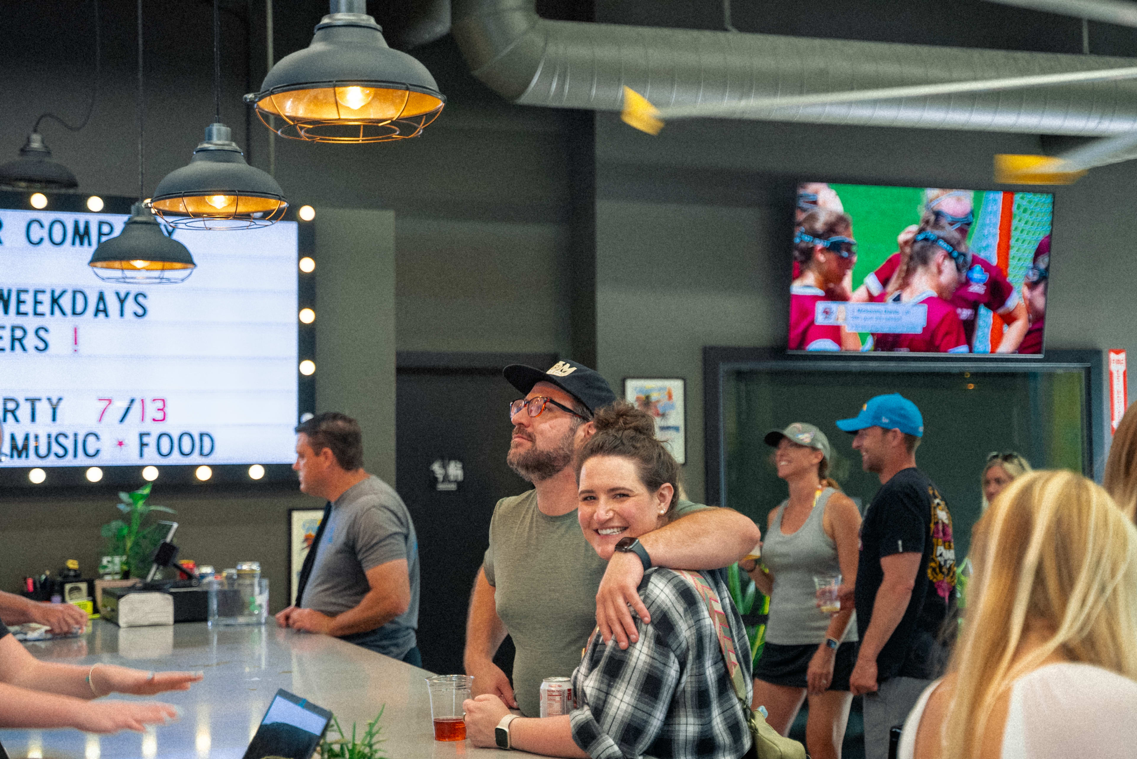 A man and a woman stand at a bar, smiling while others socialize in a lively indoor setting with screens displaying sports.