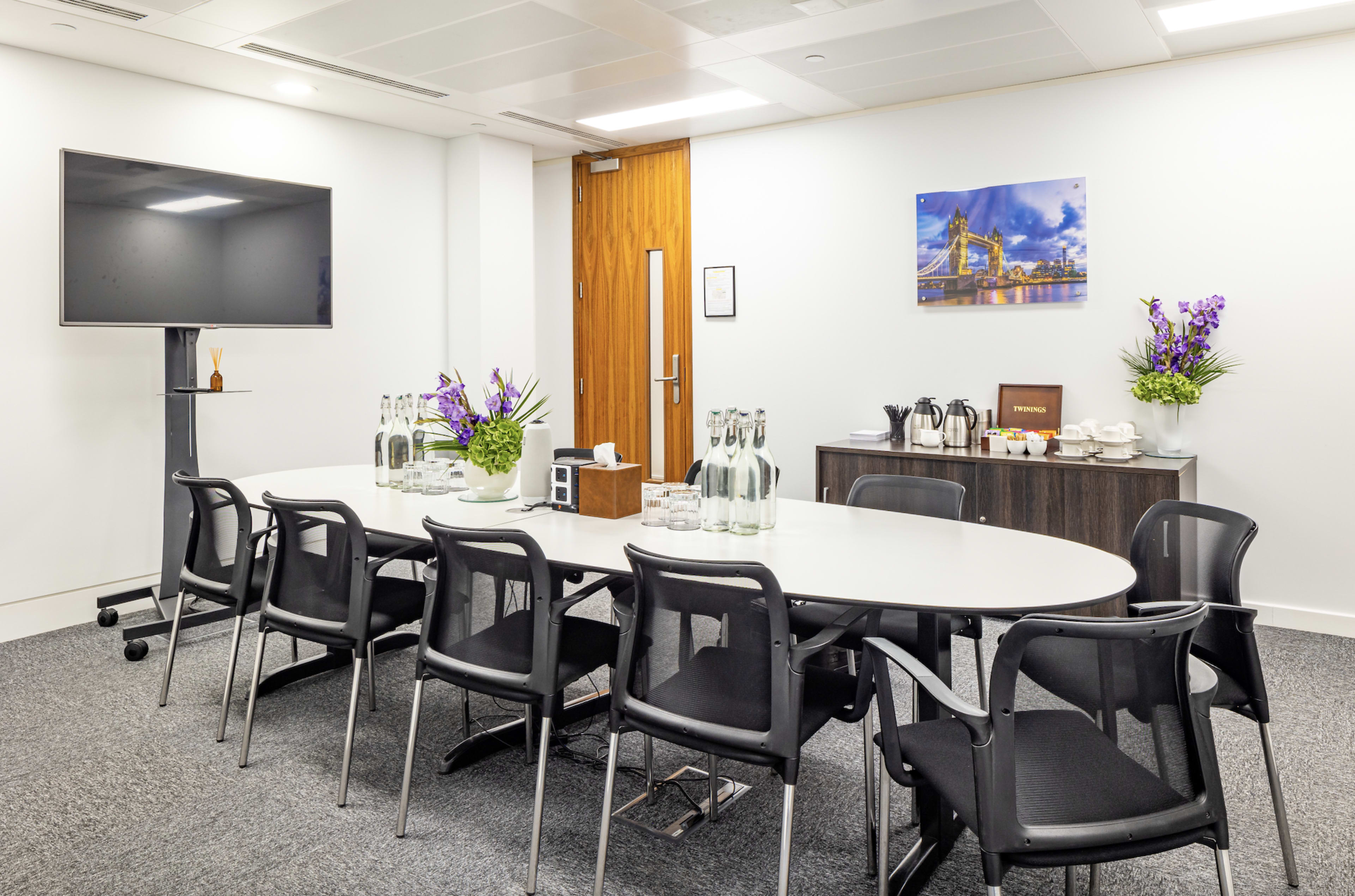 A modern conference room features a long oval table surrounded by black chairs, with a television mounted on the wall and refreshments set on a side cabinet.